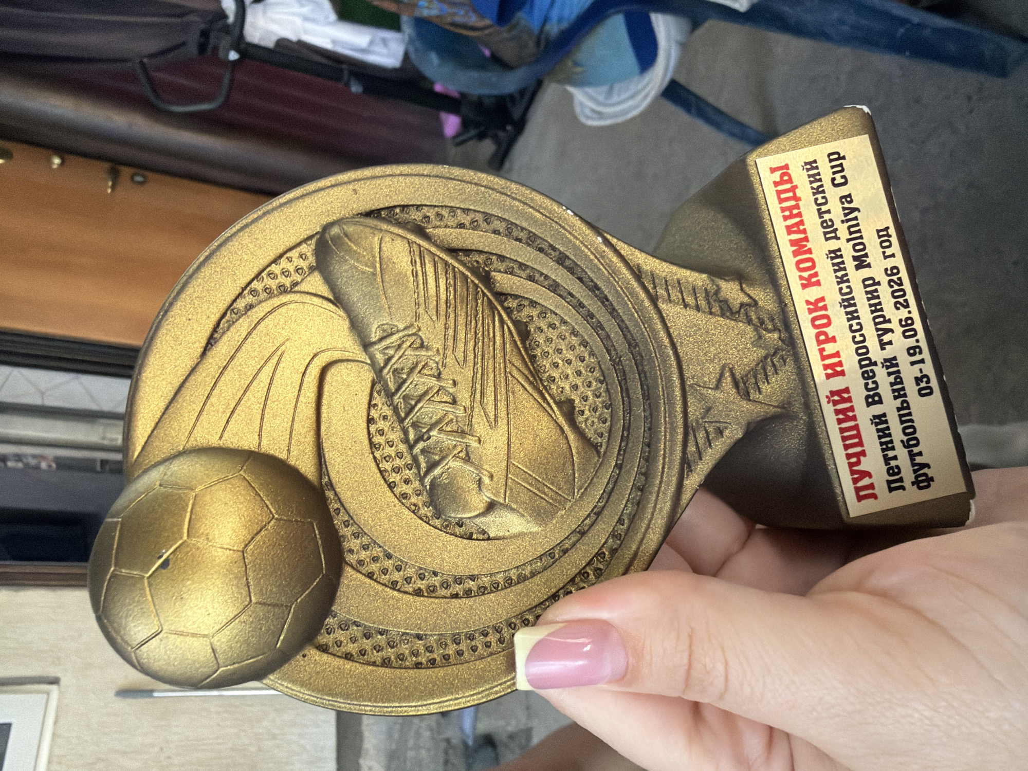 The image shows a close-up of a golden soccer trophy held by a person's hand. The trophy is ornate, featuring a soccer ball on the left and a soccer shoe on the right, set against a backdrop of swirling patterns and a star. A red and yellow label is affixed to the trophy, bearing text in Russian. The visible text reads "ЛУЧШИЙ ИГРОК КОМАНДЫ" (Best Player of the Team), "Летний Всероссийский детский футбольный турнир Molniya Cup" (Summer All-Russian Children's Football Tournament Molniya Cup), and "03-19.06.2026 год" (03-19.06.2026 year). The background is blurred, but appears to be an outdoor setting with some wooden structures and possibly a glimpse of blue fabric. The lighting suggests it might be daytime. The hand holding the trophy has a light pink nail polish on one finger. The trophy is likely being presented or displayed after a soccer tournament.