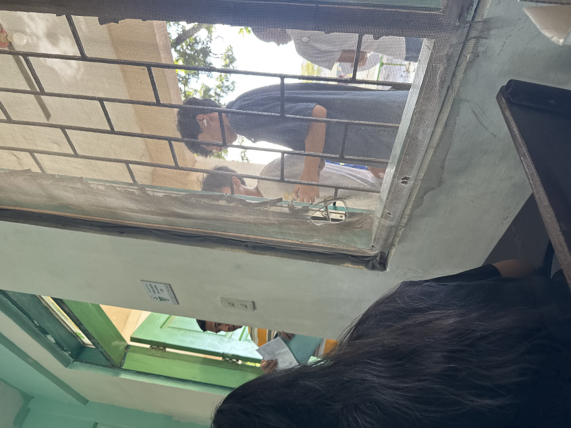 The image, taken from an indoor perspective in Davao City, Philippines, looks outwards through a window or doorway fitted with a security grill and mosquito netting. The original photograph appears to be rotated counter-clockwise.
In the foreground, at the bottom right, the back of a person's head with dark, wavy hair is visible, suggesting someone observing the scene from inside. Through the mesh screen, several individuals are present outside. A man in a dark blue or black t-shirt is prominently featured, leaning forward and peering into the opening, possibly interacting with someone inside or looking down. He has short, dark hair and an earbud in his right ear. Below him, another person in a light-colored shirt is partially visible. Further back, another individual in a light shirt is also seen.
To the left of the main window area, through an open green door or window frame, another man is visible. He is wearing a striped yellow and white shirt and is holding a stack of papers, suggesting he might be involved in an administrative process or transaction.
The interior walls are painted a light green or teal. Above the open green door, a small white rectangular sign with black text and a symbol (indiscernible due to blur) is affixed to the wall, likely an instruction or warning notice. Beyond the people outside, bright daylight illuminates trees and clear sky, indicating it is daytime with sunny weather. The presence of security bars and mosquito netting is common in the tropical climate of the Philippines.