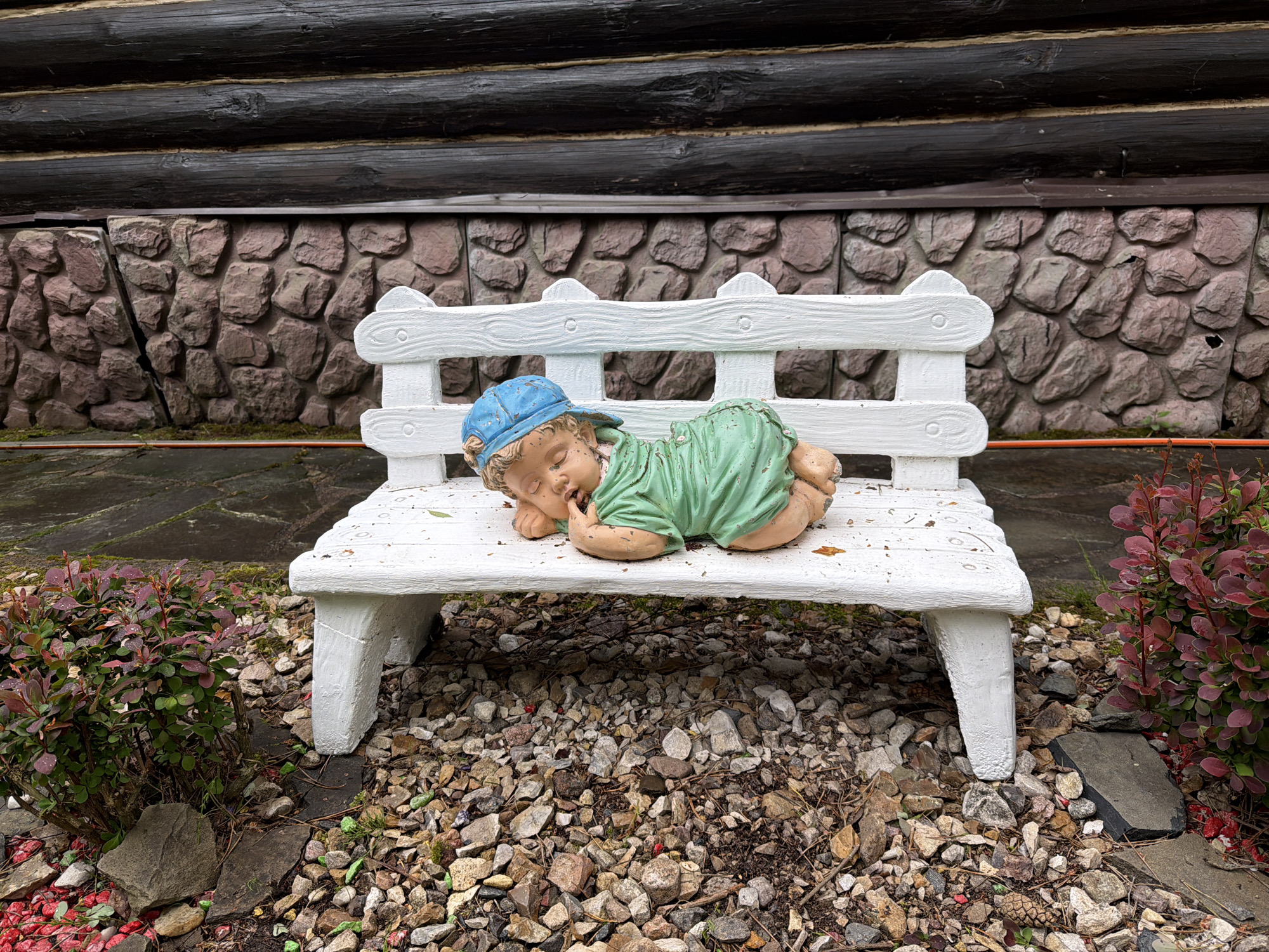 A detailed outdoor shot, likely in Zaplav'e, Russia, shows a weathered white wooden bench set on a gravel bed. Atop the bench, a statue of a sleeping child, wearing a blue cap and green outfit, is placed. The backdrop includes a stone wall and a dark wooden log structure. Bushes with green and maroon leaves flank the bench. The scene's overall condition appears weathered and naturalistic.