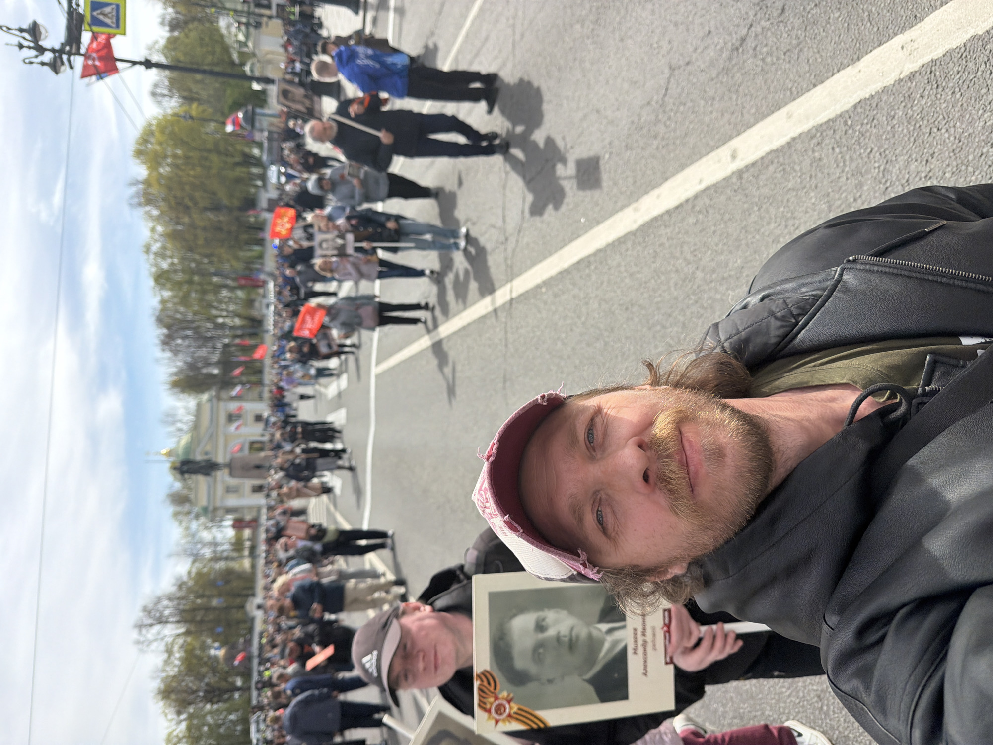 Here is a description of the image:
The photograph appears to have been taken during a public event, likely a parade or procession. A large crowd of people fills the street in the background. Many people hold portraits or banners, suggesting a memorial or commemorative event. Several red flags and banners are visible among the crowd, indicating the political nature of the event. The Winter Palace of the Hermitage Museum is visible in the background, placing the location in the city of Saint Petersburg, Russia.
In the foreground, there are two men. The man closest to the camera is smiling and wearing a baseball cap, a jacket, and he has a beard. Another man is behind the first man, also wearing a hat, and holding a portrait of a man. The portrait is framed, and a ribbon is attached to the frame.