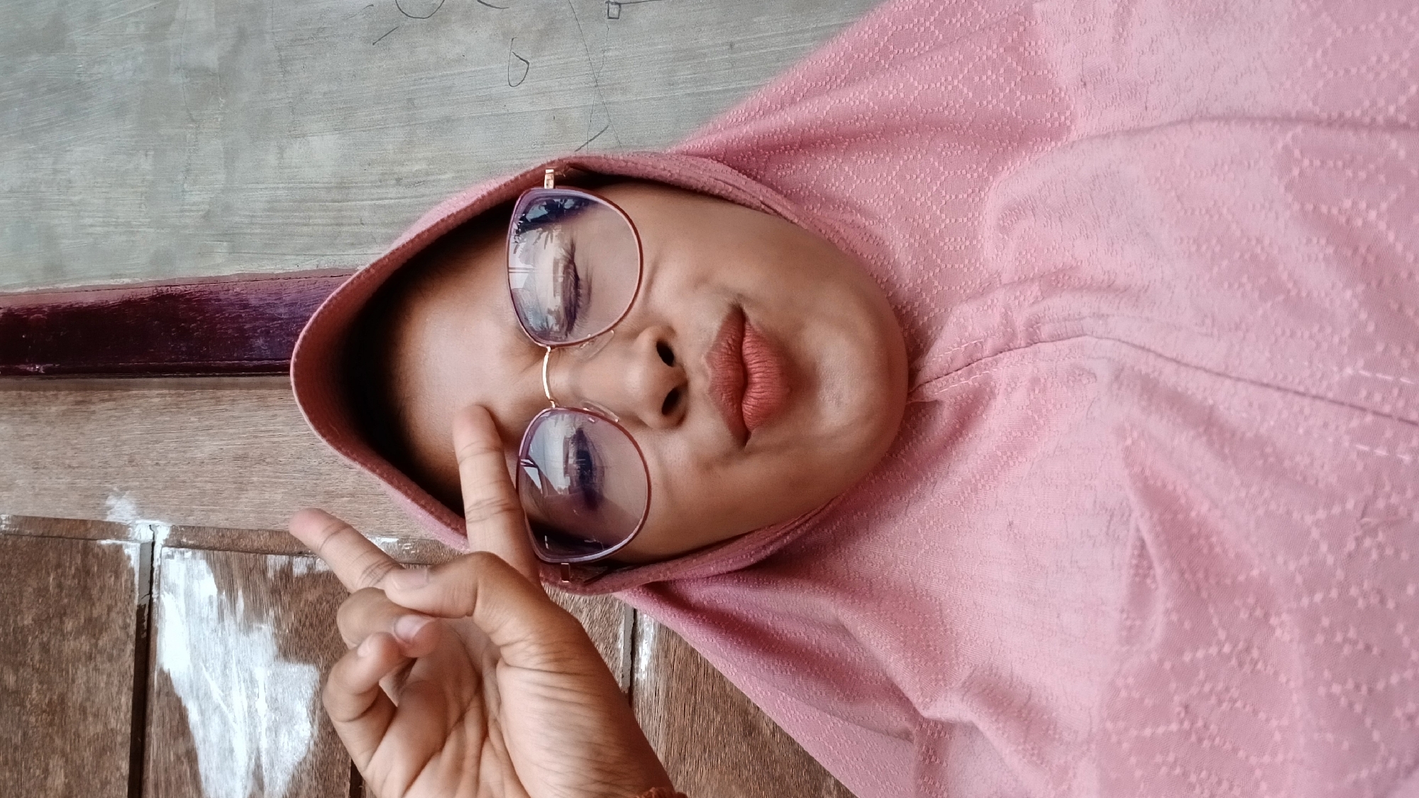 A person wearing a pink headscarf and glasses is positioned in the image. They are holding up two fingers in a peace sign. The setting appears to be outdoors, potentially near a wooden surface with traces of paint. The image lacks sufficient detail to confirm the exact location in Lhokseumawe, Indonesia.