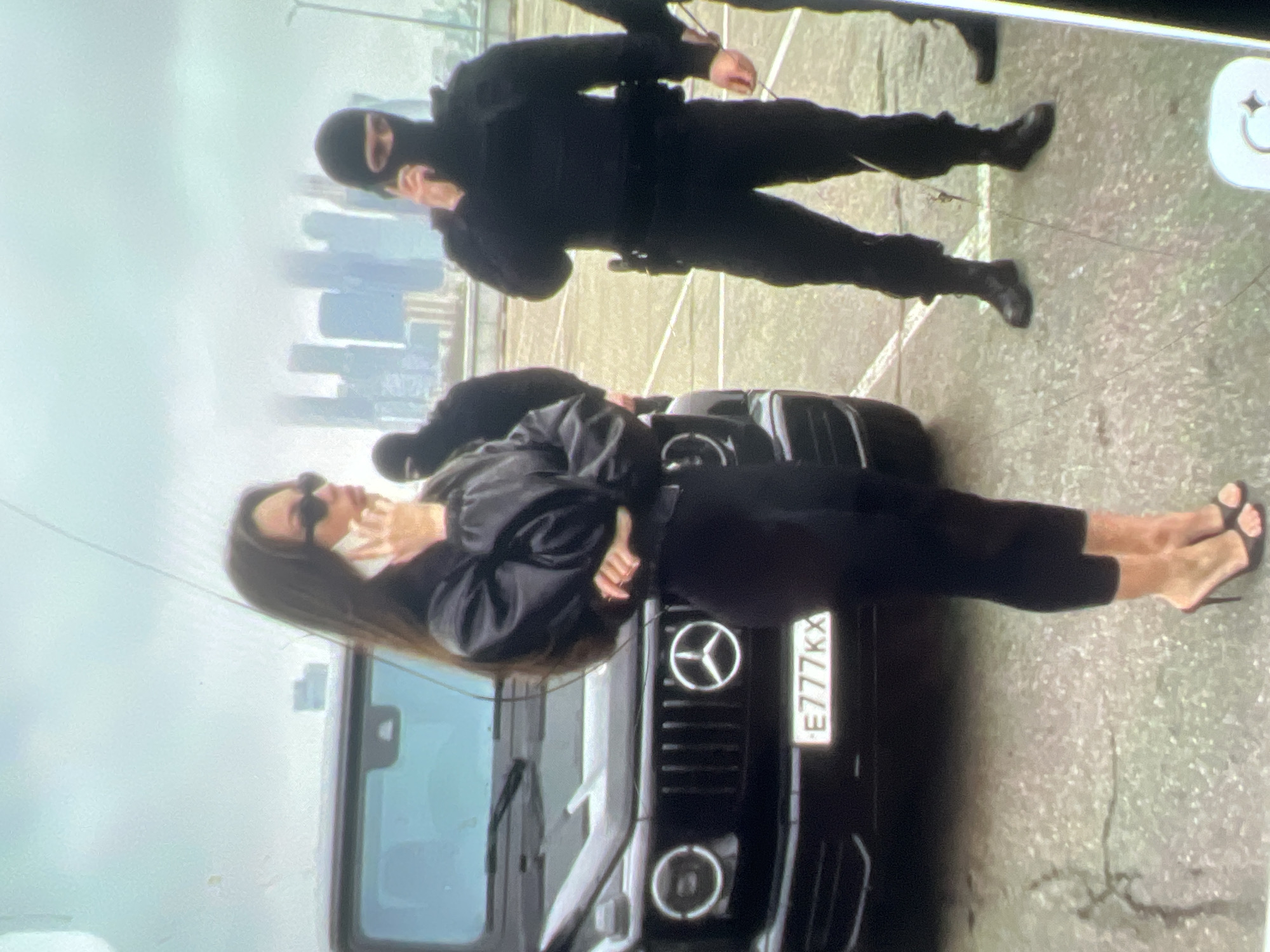 The image captures an outdoor scene in an urban environment, likely a parking lot or open area with a city skyline visible in the background under an overcast sky. In the foreground, a woman with long dark hair is standing next to a black Mercedes-Benz G-Class SUV. She is wearing a black leather jacket, black leggings, and high-heeled mules, and is holding a white smartphone to her ear. Her pose suggests she is engaged in a phone conversation. Behind the woman and the vehicle, two individuals in black tactical gear and balaclavas are visible. One is standing to the right of the woman, also appearing to be on a phone call and wearing a tactical vest and utility belt with a holster. Another individual in similar attire is partially visible to the left, behind the woman. The environment appears to be an elevated area, possibly a rooftop parking lot, with a glimpse of a city's modern architecture in the distance. The ground is paved with visible cracks.