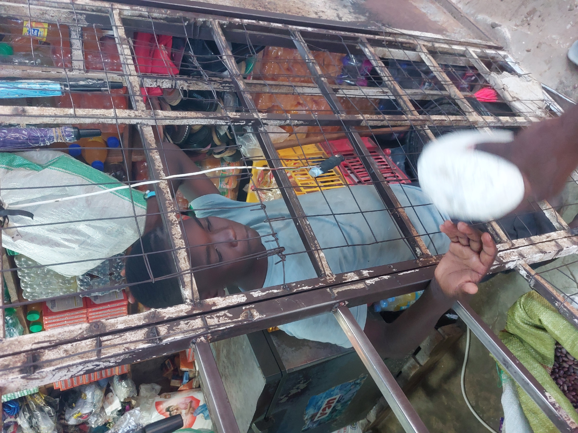 The image presents an overhead view looking down into what appears to be a market stall or small shop in Dar es-Salaam, Tanzania, through a rusty, dark metal grate.
The main subject is a person, likely male, with dark skin, resting or sleeping beneath the grate. He is wearing a light-colored (possibly blue or grey) t-shirt, and his eyes are closed, suggesting peaceful repose. His head and upper torso are visible through the grate's squares.
Beneath the metal framework, a dense collection of goods is stored. These include plastic bottles containing various liquids (some clear water, others amber-colored beverages), folded umbrellas (one blue, one purple), and red plastic crates. A large white woven sack with green stripes, possibly filled with produce like peanuts, is visible on the left side. Also visible are stacks of small, rectangular boxes, possibly cigarettes or mosquito coils, and various shoes or sandals. Towards the bottom, a cooler or display unit bears the letters "USA." A white electrical cord runs horizontally across the frame.
On the far right, a dark-skinned hand is seen holding a blurry, white object, which could be a piece of food or a small container. The overall setting suggests a bustling, utilitarian market environment. The time of day and weather are not discernible. No other specific text is clearly legible beyond "USA."