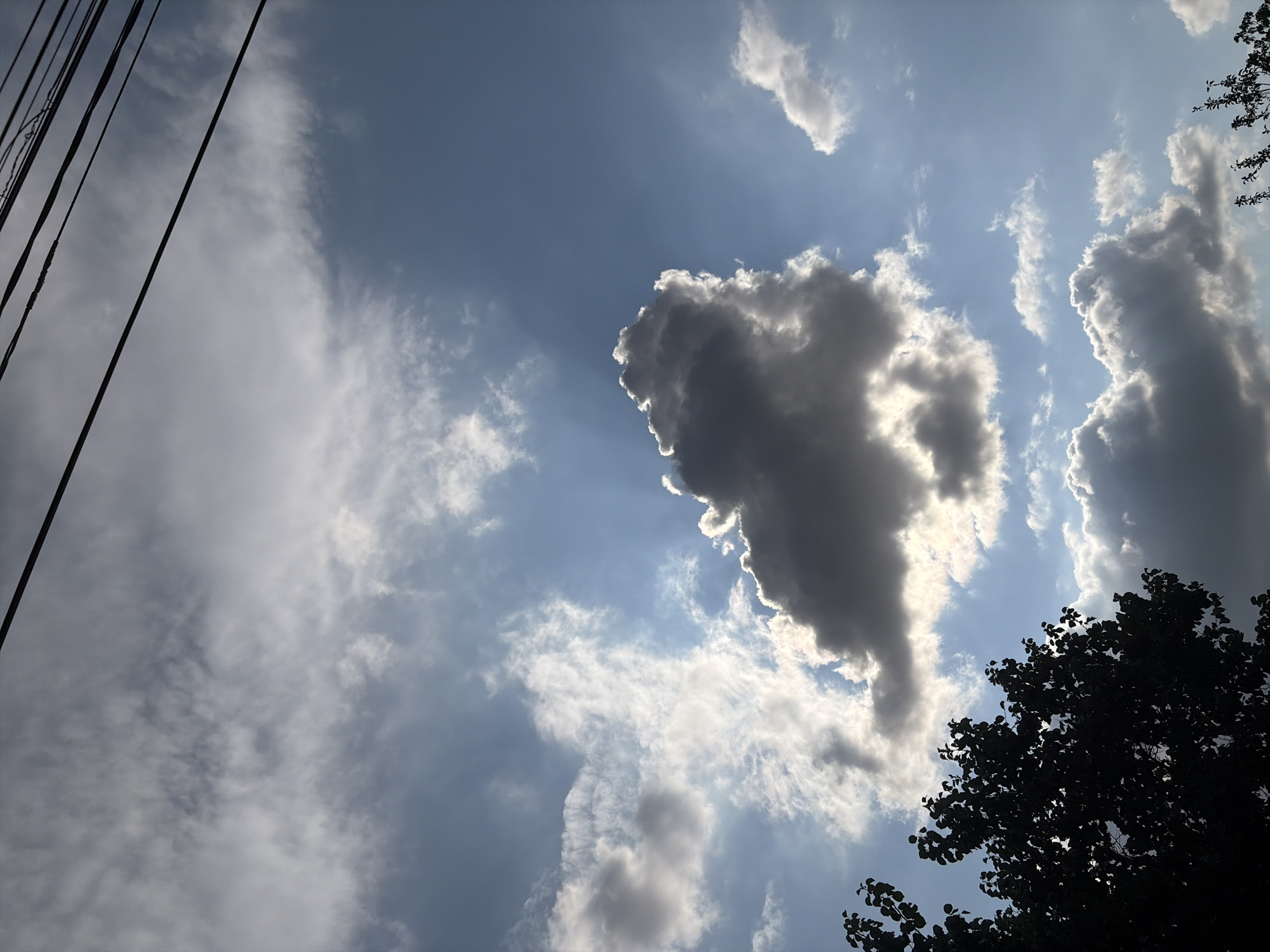 The image depicts a sky with scattered clouds and overhead power lines. A silhouette of tree branches is visible in the lower left corner. The sky is blue, with some areas of lighter and darker clouds, suggesting varying degrees of sunlight penetration. No people or specific city landmarks are identifiable, making it impossible to confirm the location in Oxentea, Moldova.
