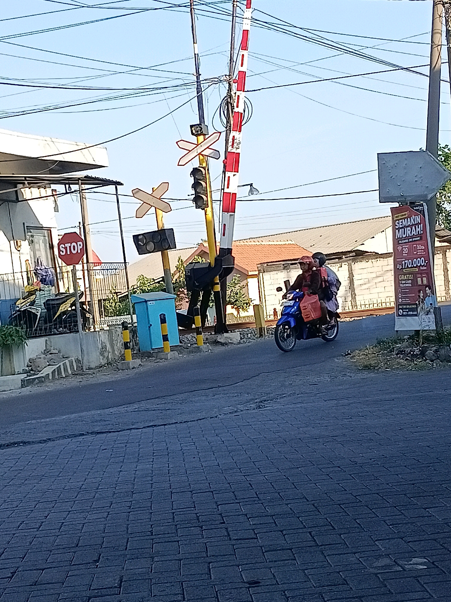 In Kletek, Indonesia, on a sunny day with a clear blue sky, a railroad crossing is depicted. A red and white striped gate arm is lowered, indicating an approaching train. A traffic light signal with red and yellow lights is visible. A motorcycle carrying two people is approaching the crossing from the right. The person in front is wearing a red helmet and a dark jacket, while the passenger behind is wearing a dark jacket and a light-colored scarf. They have red bags attached to the back of the motorcycle. A "STOP" sign is posted on the left side of the road. Buildings with orange roofs and white walls are in the background, along with overhead power lines. A blue utility box with yellow and black striped poles is situated on the side of the road. The foreground shows a paved road with brick-like patterns. A billboard advertising "SEMAKIN MURAH!" with prices and promotional offers is visible on the right.