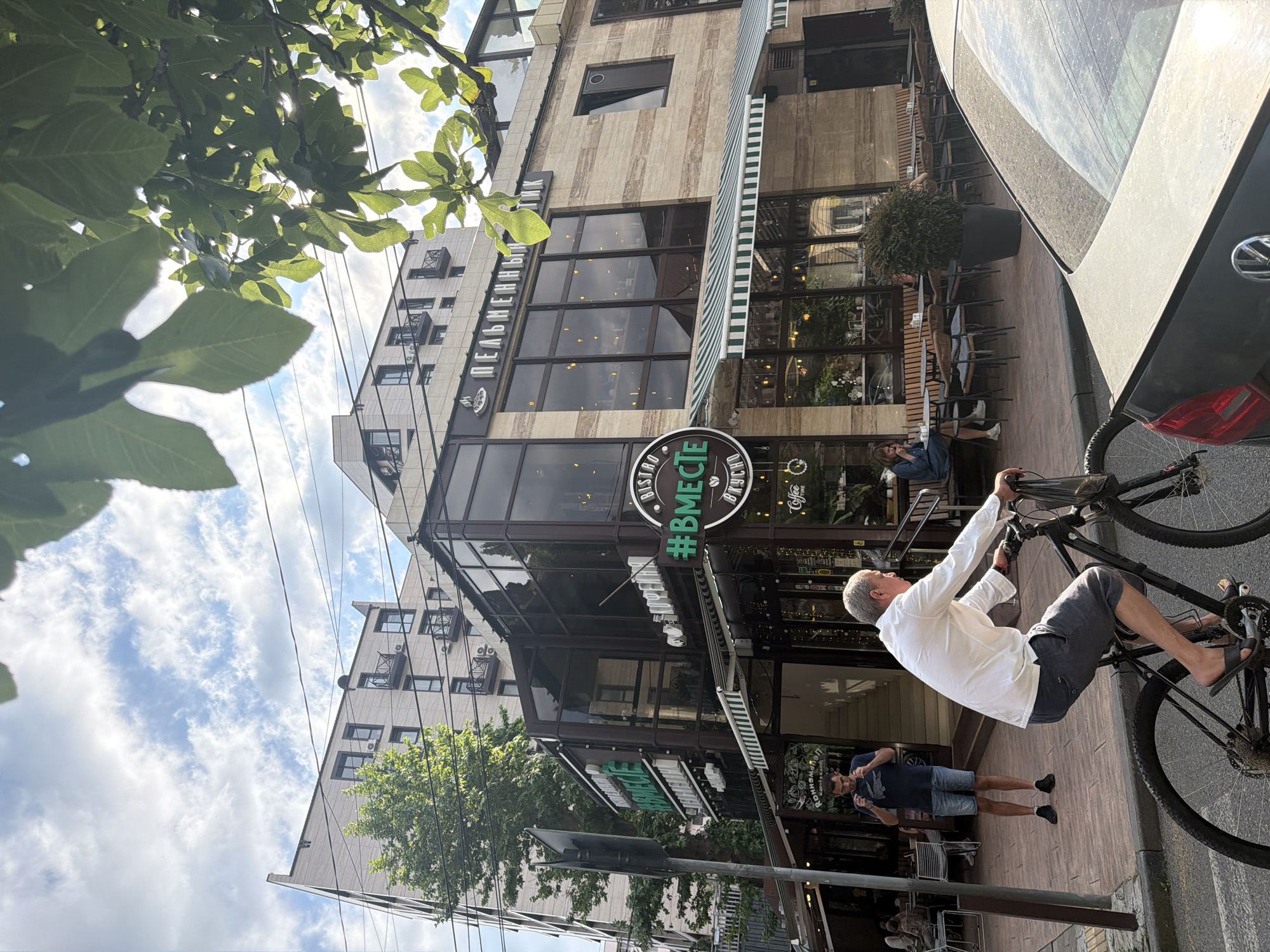 A man in a white shirt and grey shorts is riding a bicycle on a street in Sochi, Russia. He is on a sidewalk next to a building with large windows and awnings. A car is parked on the right side of the image, and another man in a blue shirt and shorts is standing on the sidewalk in the background, looking at his phone.
The building appears to be a restaurant or cafe, with signs visible that read "#ВМЕСТЕ" (which translates to "#TOGETHER") and "ПЕЛЬМЕННАЯ" (which translates to "Dumpling House"). The time of day appears to be afternoon, with sunlight casting shadows on the street. The weather seems pleasant, with a partly cloudy sky. The scene is urban, with trees and buildings forming the backdrop. The man on the bicycle is the primary focus, and his posture suggests he is actively riding. The other individuals are secondary subjects, engaged in their own activities.