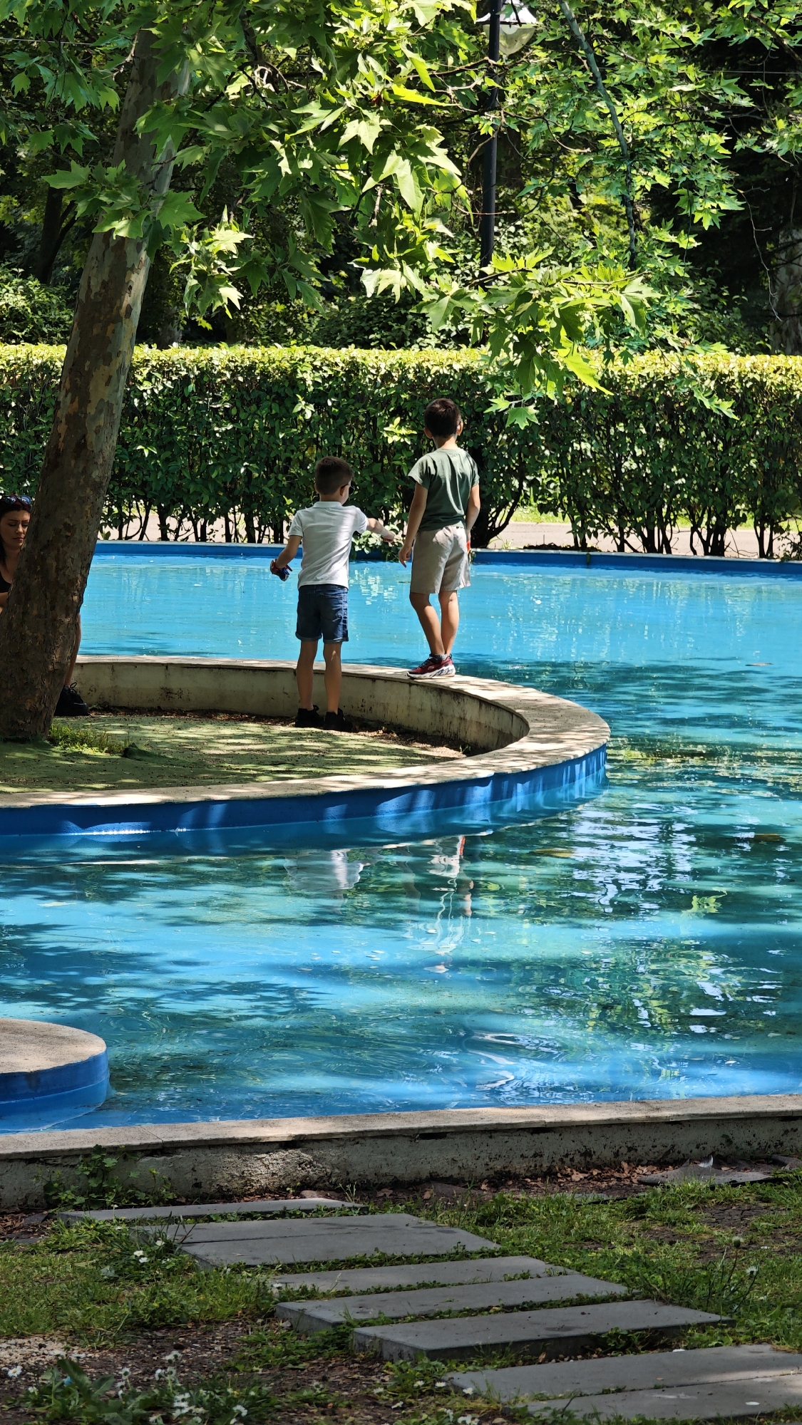 In a daytime, outdoor scene likely in Burgas, Bulgaria, two young boys are standing on the edge of a semi-circular, elevated structure adjacent to a turquoise-colored body of water, possibly a pool or ornamental pond. The boy on the left wears a white shirt and dark shorts, and is holding an object in his hand. The boy on the right is clad in a green shirt and beige shorts. The boys are facing the water, their attention drawn to something within or on it. Behind them, a hedge forms a barrier, with trees providing a backdrop. To the left, a tree trunk stands partially obscuring a person seated nearby. In the foreground, a path constructed of rectangular stones leads towards the structure with the water.