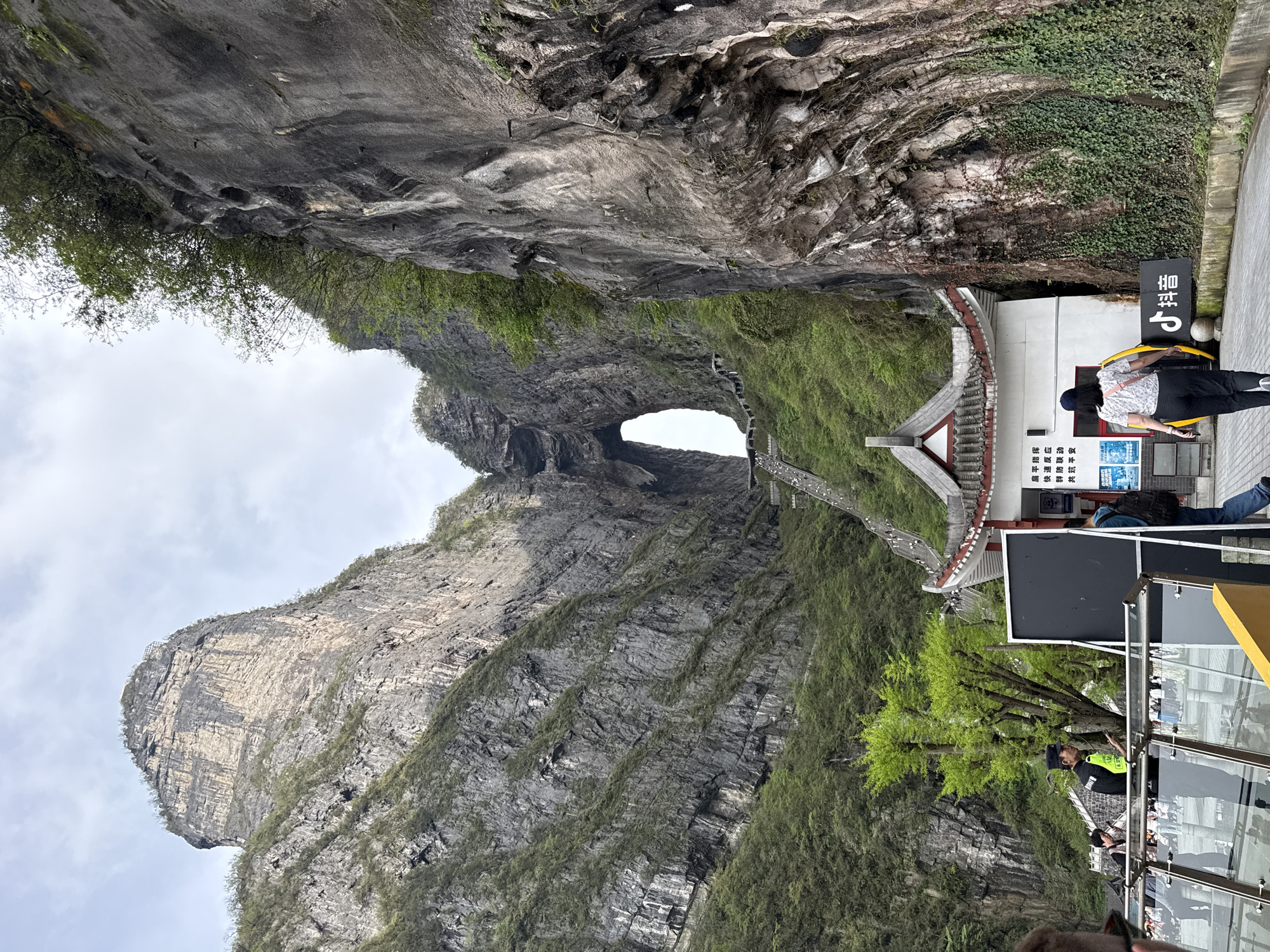 This photo depicts Tianmen Cave, also known as Heaven's Gate, in Tianmen Mountain National Park in Zhangjiajie, China. The large natural archway is visible in the mountain face, with a staircase leading up to it. To the right of the staircase, there is a building with a traditional Chinese-style roof. There are people visible near the building. A partially visible sign and viewing platform are in the lower right. The day appears overcast.