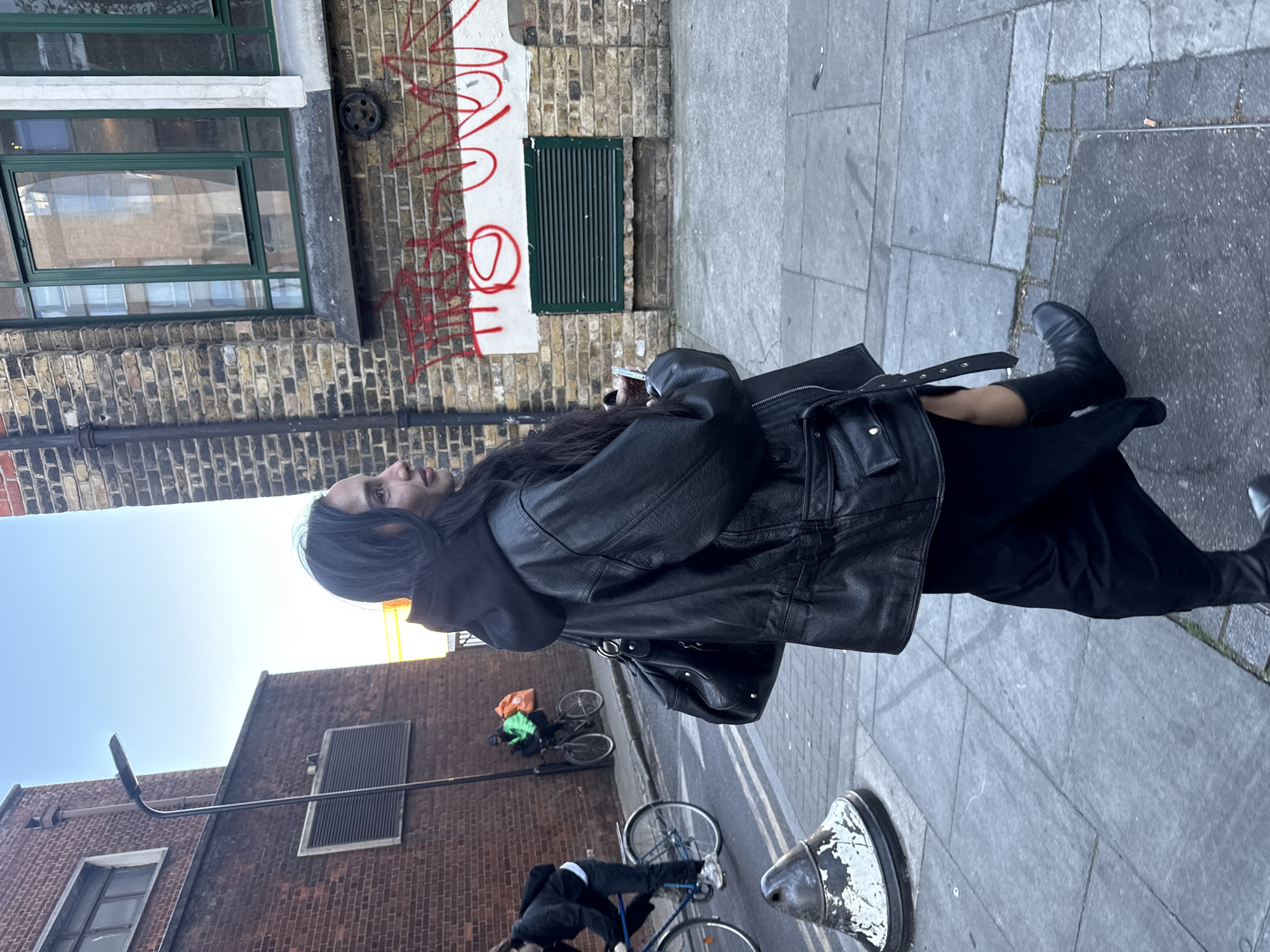 The image captures an outdoor street scene in London, featuring a person standing on a grey flagstone sidewalk next to a brick building. The main subject, with long dark hair, is positioned in the mid-ground, facing slightly to their left and looking upwards. They are dressed in a black leather jacket over a black hooded top, black trousers, and black boots. They hold a smartphone in their right hand near their chest and carry a black shoulder bag on their left.
The setting consists of an older brick building on the left, featuring multiple windows, some with green frames, and a green slatted vent. A section of a white wall on this building is covered with illegible red graffiti. Further in the background, another brick building is visible. A clear blue sky is seen above the buildings.
In the mid-ground, a black metal pole stands. On the street behind the main subject, two individuals are riding bicycles. One cyclist wears a bright green jacket and carries a distinctive orange delivery bag on their back, while the other cyclist is partially obscured. A distressed white and black bollard is visible in the lower right foreground.
The scene appears to take place during a clear daytime, with bright, even lighting. The overall impression is that of a typical urban environment. The main subject's expression is neutral as they gaze upwards.