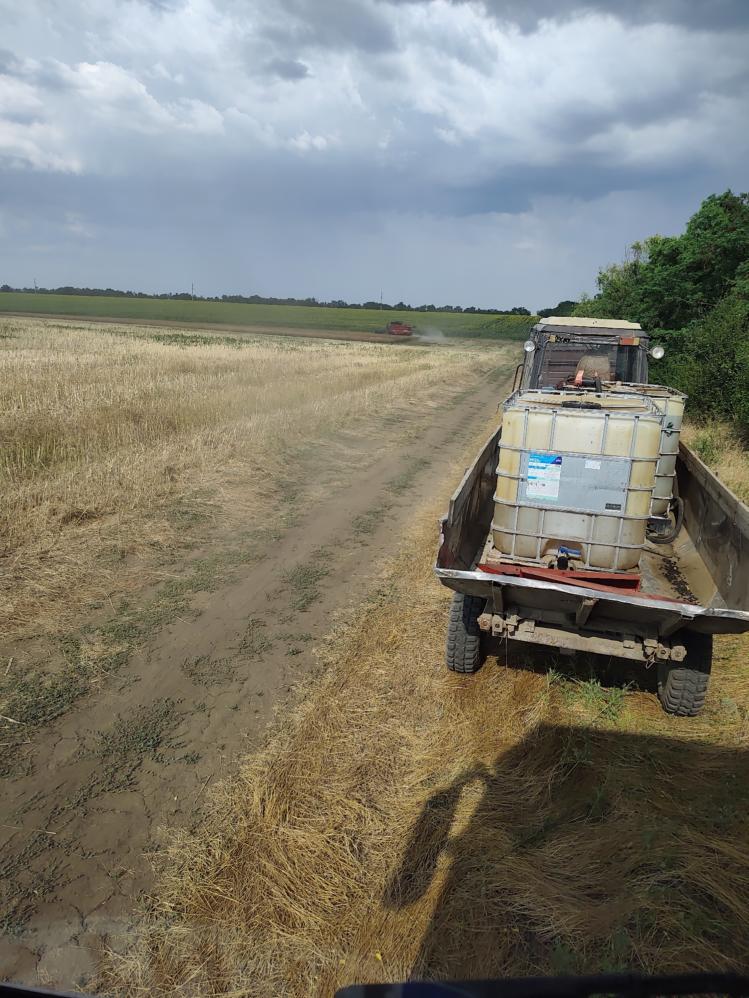 In a rural setting, likely in Alexandru Ioan Cuza, Moldova, a trailer is being pulled by a tractor through a field of dry, yellow stubble. The trailer carries two large white plastic containers filled with liquid, likely agricultural chemicals, as indicated by a label on one of the containers. The trailer is attached to a tractor, the front of which is visible in the foreground. In the background, another red tractor is working in a green field under a cloudy sky. The overall environment appears to be agricultural, possibly during harvest season. The scene is captured from a perspective slightly above and behind the trailer.