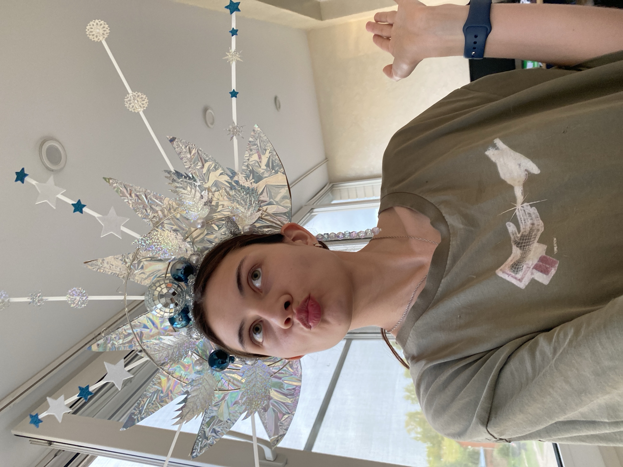 A young woman with her lips pursed and eyes looking upwards is wearing a large, elaborate headdress adorned with shiny silver elements, blue ornaments, and small disco balls. She is also wearing a green t-shirt with a graphic print and a silver necklace. A dark blue smartwatch is visible on her left wrist. Her right hand is raised, palm facing forward. The background suggests an indoor setting with a window overlooking a green, outdoor area, possibly a residential or semi-rural environment in Votkinsk, Russia.
