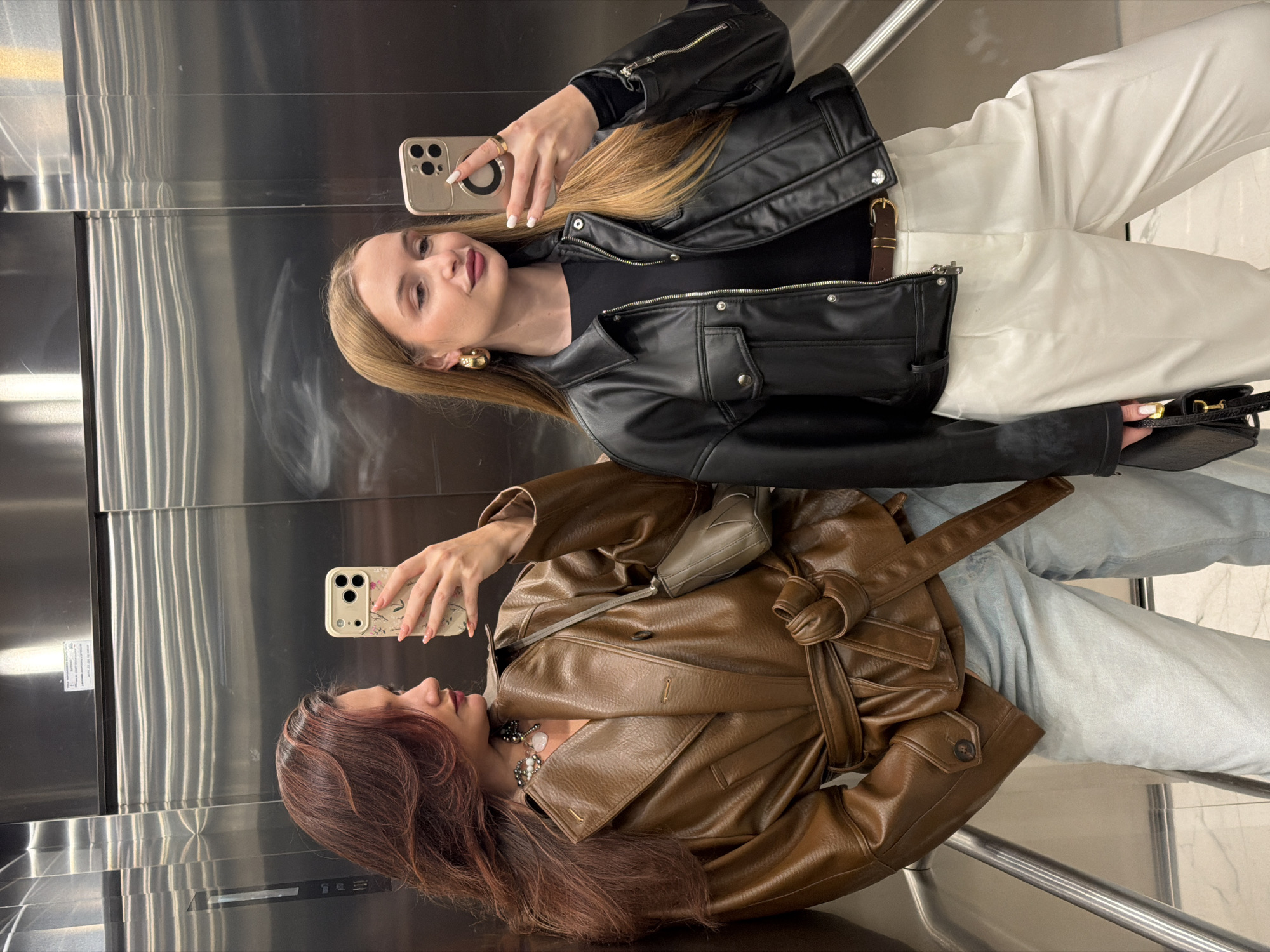 Two young women are taking a selfie in an elevator with a polished metal interior. The woman on the top is blonde and wears a black leather jacket, a black shirt, and white pants. She holds a smartphone with a gold case. The woman on the bottom has reddish-brown hair and wears a brown leather trench coat over light blue jeans. She also holds a smartphone. Both women are looking at their phones, presumable taking the selfie. The elevator has metal handrails. The location appears to be a modern building, but there is no indication of the specific city or country.
