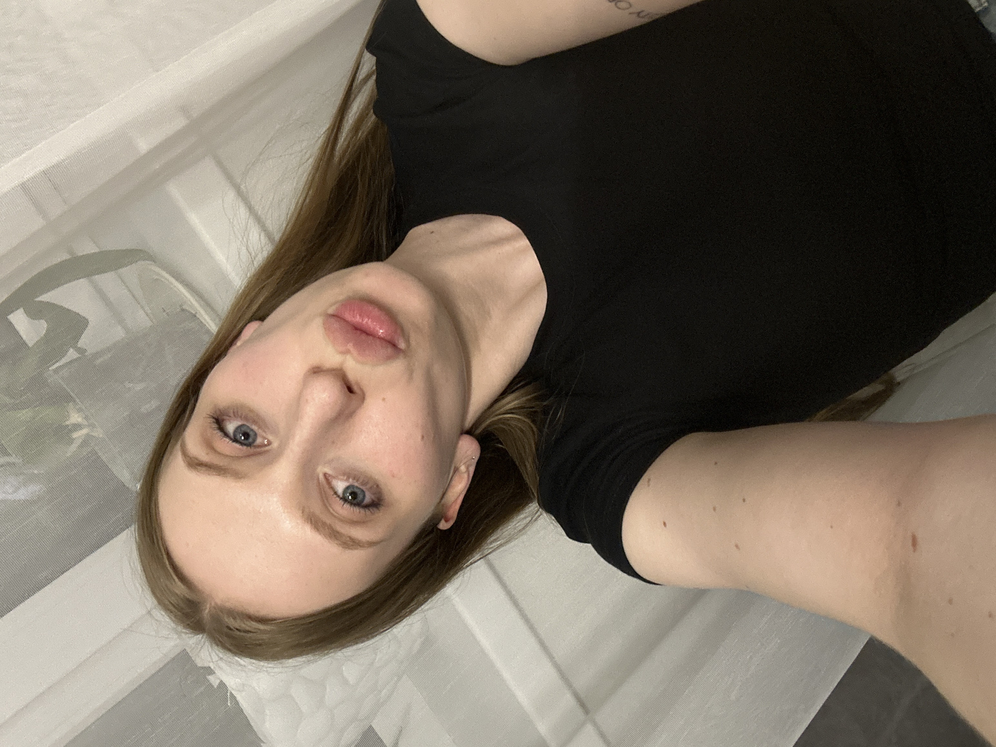 A selfie of a woman with fair skin and blue eyes. She has long, light brown hair. She is wearing a black T-shirt. The background appears to be a white surface, possibly a bed or another fabric. The location is unknown, but the query includes Chaikovsky, Russia.