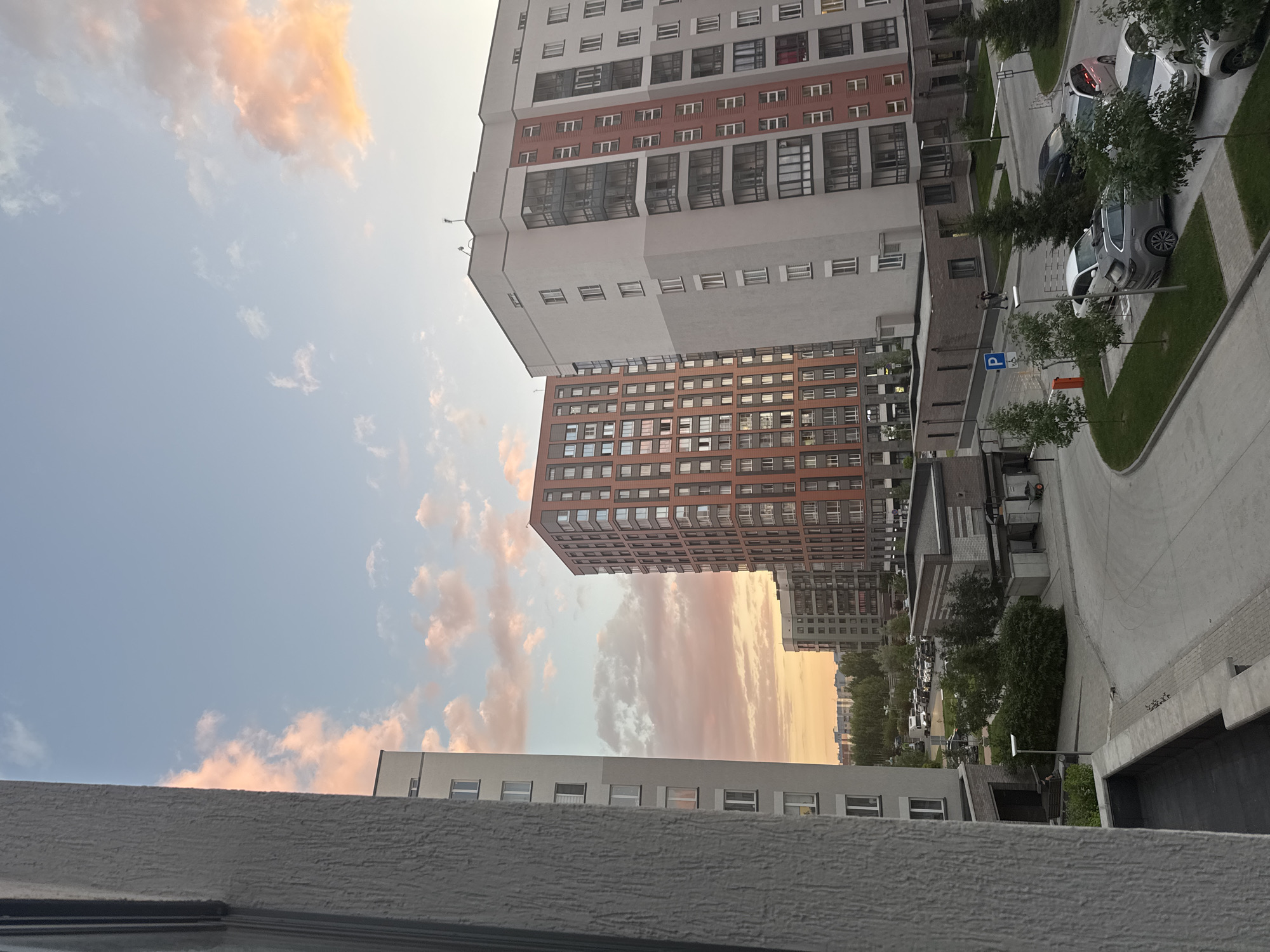 This media file depicts a view of modern residential buildings in Koltsovo, Russia. The image is oriented vertically, showing multiple apartment blocks of varying heights and architectural styles. The buildings are predominantly light-colored with darker brick accents and numerous windows. In the foreground, a textured concrete ledge is visible, suggesting the image was taken from a balcony or window.
The scene includes a paved area with parked cars, lined with small trees and green lawns. A blue parking sign with a white "P" is visible. The sky is a soft blue, with scattered clouds tinged with orange and pink, indicating either sunrise or sunset. The lighting suggests it is late afternoon or early evening.
A few indistinct figures can be seen walking on the paved area between the buildings and cars. The overall atmosphere is calm and residential. The location is explicitly stated as Koltsovo, Russia. There are no discernible emotions conveyed, and the image focuses on the urban landscape. No visible text other than the parking sign.