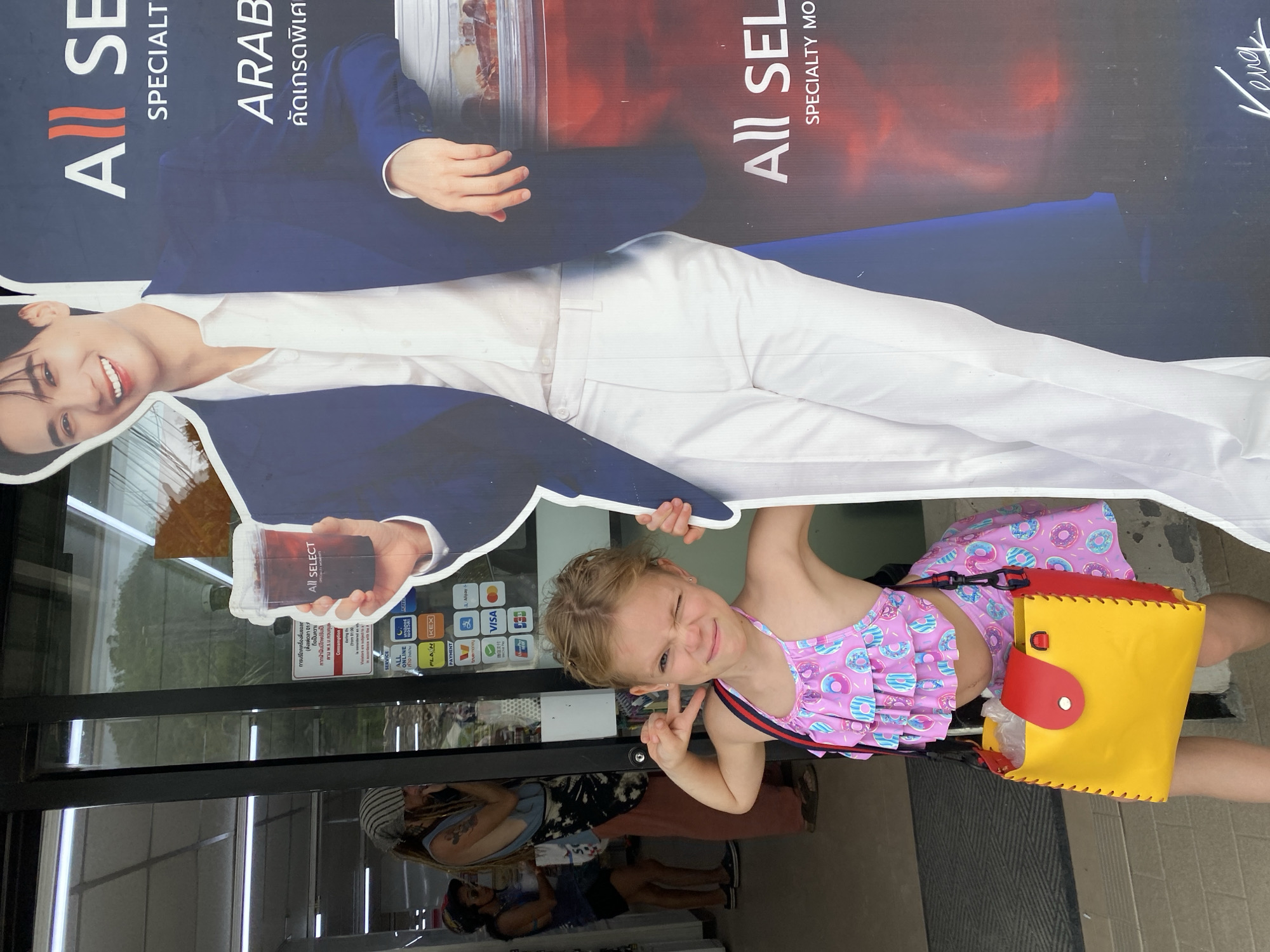In the city of Ko Pha Ngan, Thailand, a young girl with blonde hair is posing playfully in front of a life-size cardboard cutout of a smiling man. The girl is wearing a pink swimsuit with a donut pattern and a yellow and red purse. The cutout features the man in a blue suit and white shirt, holding a cup of "All Select" beverage. Behind them, two women are visible through a store window, and a sign displaying payment options is attached to the store's entrance. The environment appears to be an outdoor setting, possibly a street or marketplace.