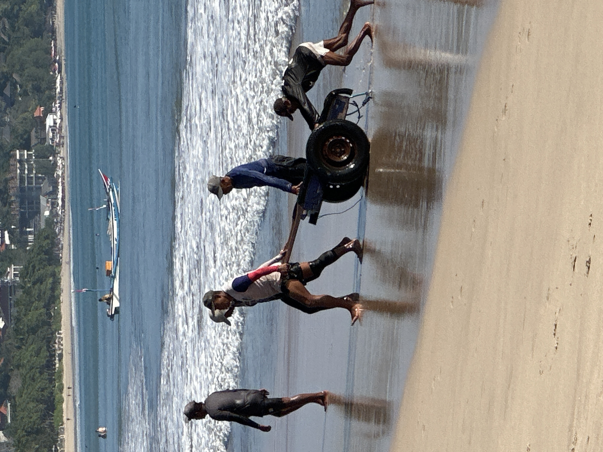 The image shows four men on a beach in Kuta, Indonesia. Two men in the foreground are pulling a cart with large wheels on the wet sand, with waves lapping at their feet. One man is pushing the cart from behind, and another man is pulling it from the front using a wooden beam. Behind them, to the left, another man walks towards the water, and further back, two men are partially visible near the surf. In the background, a traditional outrigger boat with a red and white flag is visible in the ocean, with buildings and lush vegetation lining the coastline. The sky is clear, and the sun is shining.