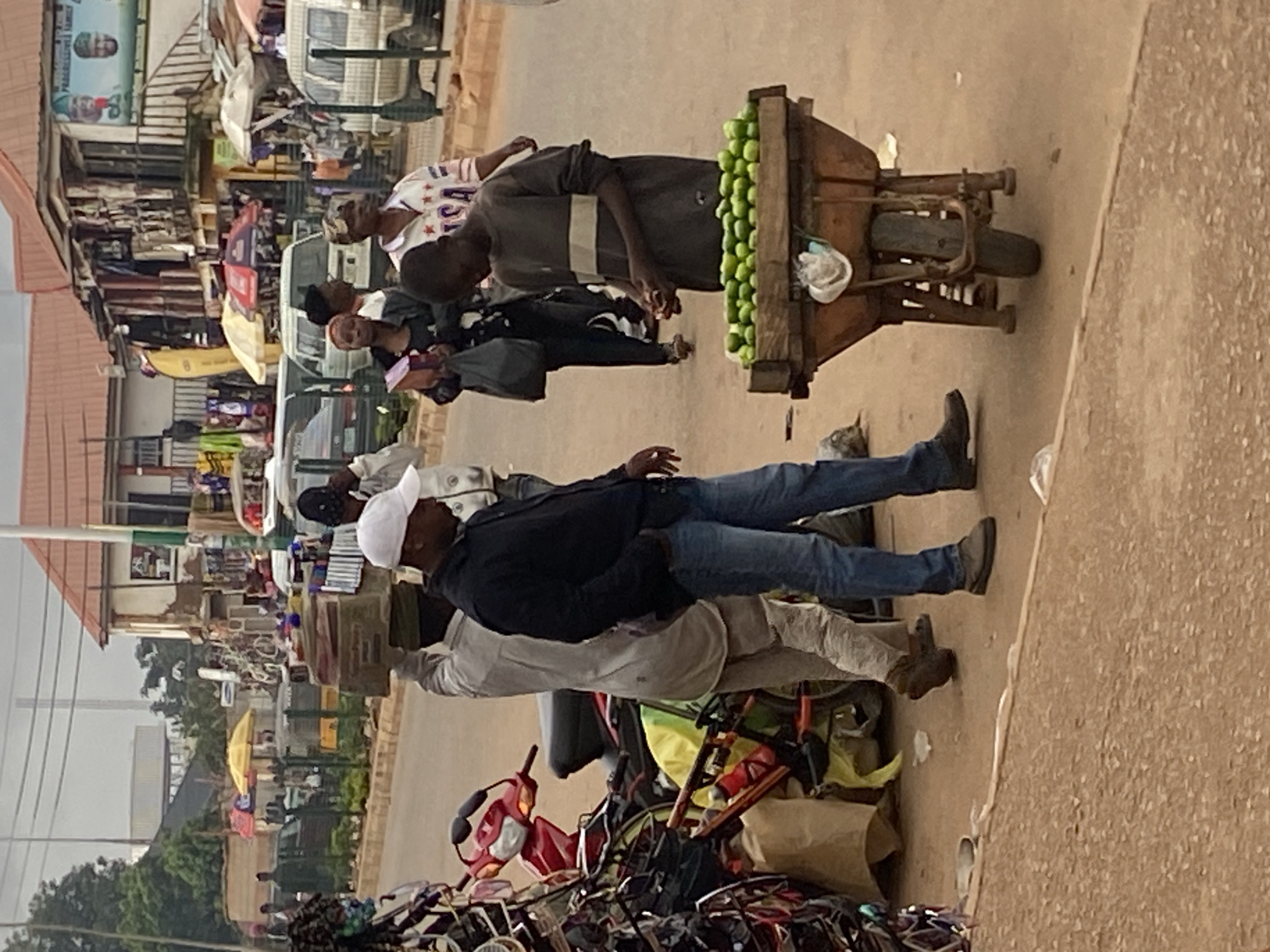 The image depicts a street scene in Bukuru, Nigeria, characterized by a bustling marketplace environment. In the foreground, a young boy is tending to a wheelbarrow filled with green produce, likely limes, positioned on a dusty street. To his left, a man wearing a white cap and blue jeans is walking past, his hands in his pockets, suggesting a casual interaction or observation. Behind them, another person is carrying a large box on their head, a common sight in Nigerian street markets.
The background reveals a row of shops and stalls, some with goods displayed outside. Several vehicles, including vans and motorcycles, are present, indicating transportation and activity. Umbrellas and awnings provide shade, typical for a market setting. Power lines are visible in the upper portion of the image, typical of urban infrastructure. The overall impression is one of everyday life and commerce in a Nigerian city.