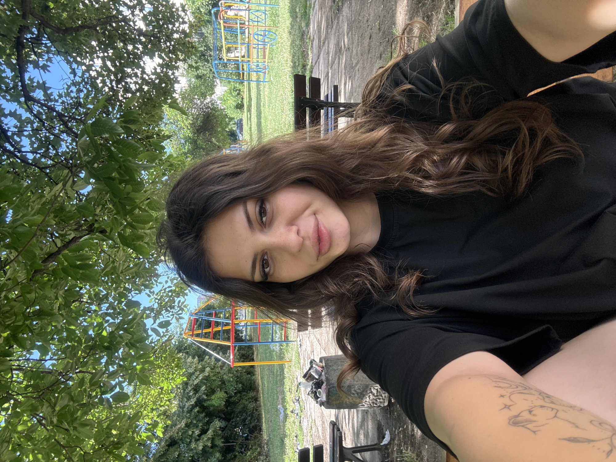 An outdoor, medium-shot, eye-level selfie is taken by a young woman with long, dark brown, wavy hair. She is wearing a black t-shirt and has a tattoo on her left arm. Her expression is neutral, with her lips slightly parted.
The background suggests a park or playground setting. In the middle ground, there is a concrete bin filled with trash, and to the left and right, park benches are visible. Behind the benches, a playground structure with red, yellow, and blue bars is situated on a grassy area. Further back, another playground structure, resembling a stylized bus, is also present. The environment is characterized by lush green trees providing shade, with a bright blue sky visible through the leaves. The overall scene appears to be a public outdoor space, possibly in the city of Plovdiv, Bulgaria, given the context provided.