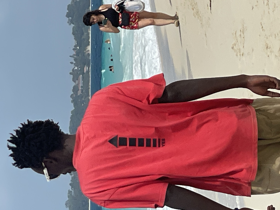 The image captures a sunny beach scene in Zanzibar, Tanzania. In the foreground, an individual with dark skin and dark, kinky hair is seen from the back, wearing white-framed sunglasses and a loose-fitting red t-shirt. The t-shirt features a prominent black upward-pointing arrow design on the back, composed of stacked horizontal rectangles, with the word "YES" positioned at its base. The individual also wears khaki-colored lower garments.
To the right, in the midground, a female with dark hair walks on the sandy beach. She is wearing a black tank top, patterned red shorts, and a black fanny pack, while holding a light-colored bag in her left hand and looking down at a smartphone held in both hands. In the background, several individuals are visible swimming or wading in the clear, turquoise-blue ocean where white waves are breaking. The distant coastline features green, vegetated hills dotted with buildings under a clear blue sky.