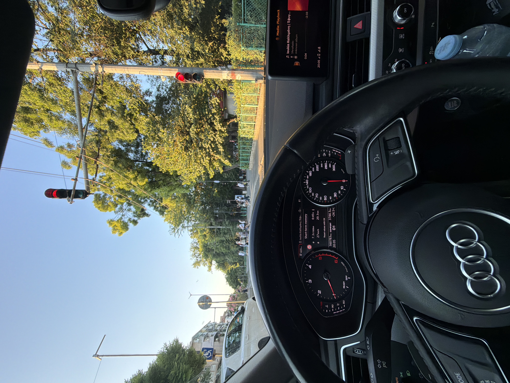 The image is taken from the interior of a car, looking out at a city street in Burgas, Bulgaria. The perspective is from the driver's seat, with the steering wheel and dashboard clearly visible. The dashboard displays speed, engine temperature, and other vehicle information. A screen shows "Media: Playback" with Greek text and album art. Outside the car, a traffic light is red, indicating a stop. Trees line the street, and several people are walking on the sidewalk. A white car is parked on the left side of the street, and buildings are visible in the background. The sky is clear and blue, suggesting a sunny day.