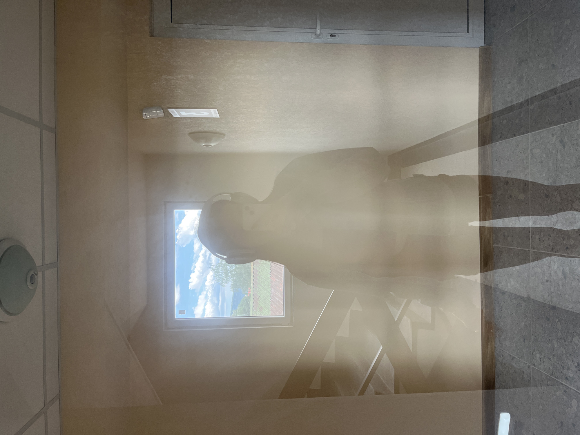 The image depicts an indoor scene with a strong, translucent reflection or superimposed figure of a person. The setting appears to be a stairwell or hallway within a building, with light-colored (beige/tan) walls and a gray and dark gray tiled floor.
In the foreground, a ghost-like, transparent figure of an individual is prominently visible, positioned as if standing or moving. This person is wearing over-ear headphones. The transparency of the figure allows the background elements to show through.
Through the translucent figure and in the background, a window is visible, showcasing a bright, sunny daytime sky with fluffy white clouds and blue patches. Outside the window, green foliage, possibly trees, and what appears to be a red-tiled roof are discernible, suggesting an outdoor landscape.
Inside the building, a set of stairs with railings can be seen, partially obscured by the superimposed figure. A circular light fixture is present on the ceiling. On a wall, a white rectangular fixture displays faint text or numbers, specifically "123," alongside another symbol resembling an 'I' or a small rectangle. Another circular, white, and greenish-blue fixture is on the left wall.
The bright natural light from the window indicates it is daytime. The overall impression is one of an ethereal presence within an otherwise ordinary, modern interior space, possibly created by a reflection on a glass surface or a double exposure photograph.
