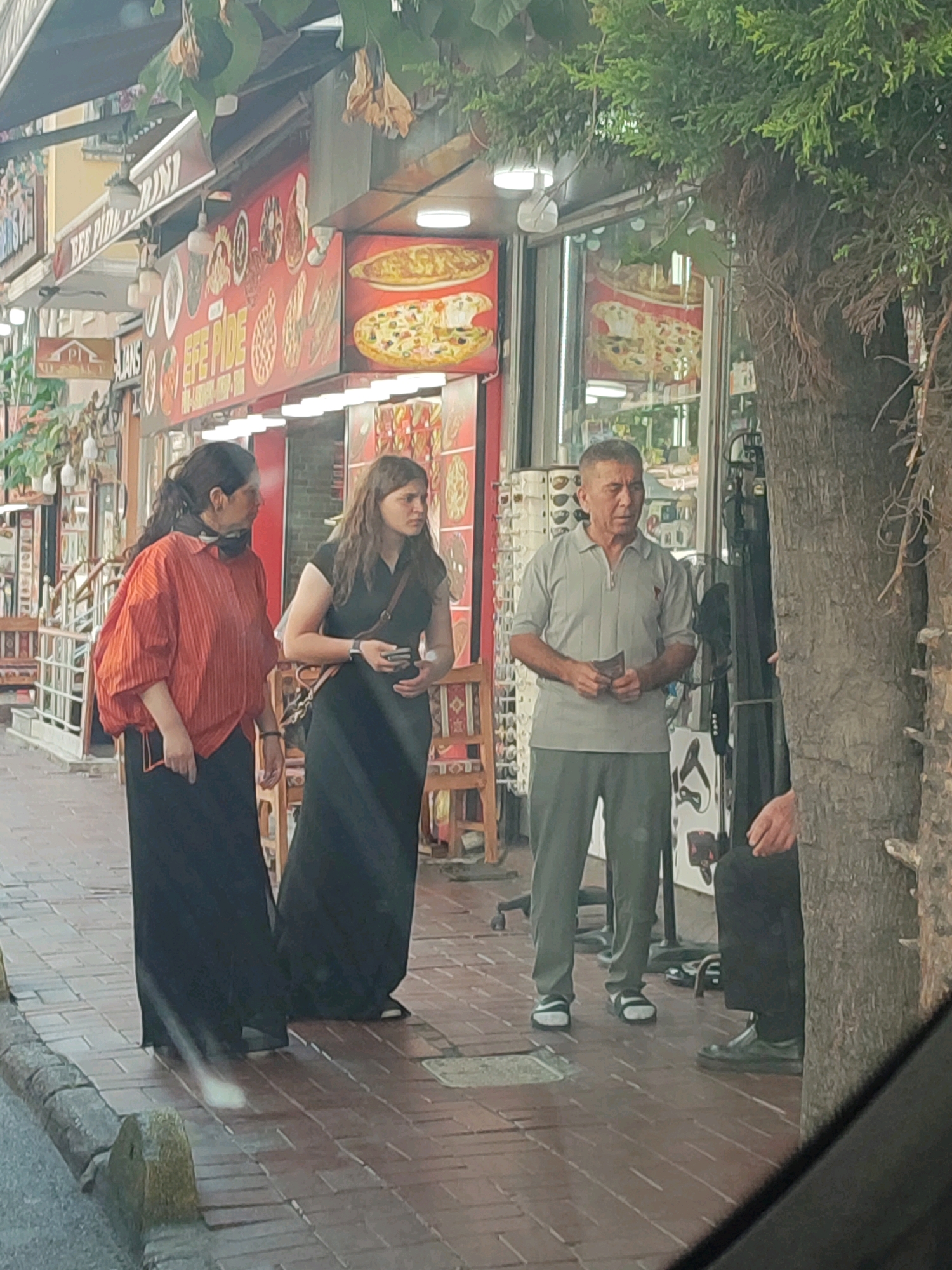 The image depicts a street scene in Istanbul, Turkey. In the foreground, three individuals are visible on a paved sidewalk. To the left, a woman wearing an orange, vertically striped shirt and a long black skirt stands with her back slightly turned, holding a small dark object in her left hand. Next to her, a younger woman in a long black dress and a crossbody bag is looking to her right and holding a smartphone. In the center of the frame, an older man in a light gray short-sleeved polo shirt and green pants is facing forward, holding a small item, possibly money, in his hands. He is standing in front of a shop with food displays. Behind the people, a shop facade is visible with various food items advertised, including pizzas and flatbreads. A large tree trunk is prominent on the right side of the image. The environment suggests a commercial street with pedestrian traffic.