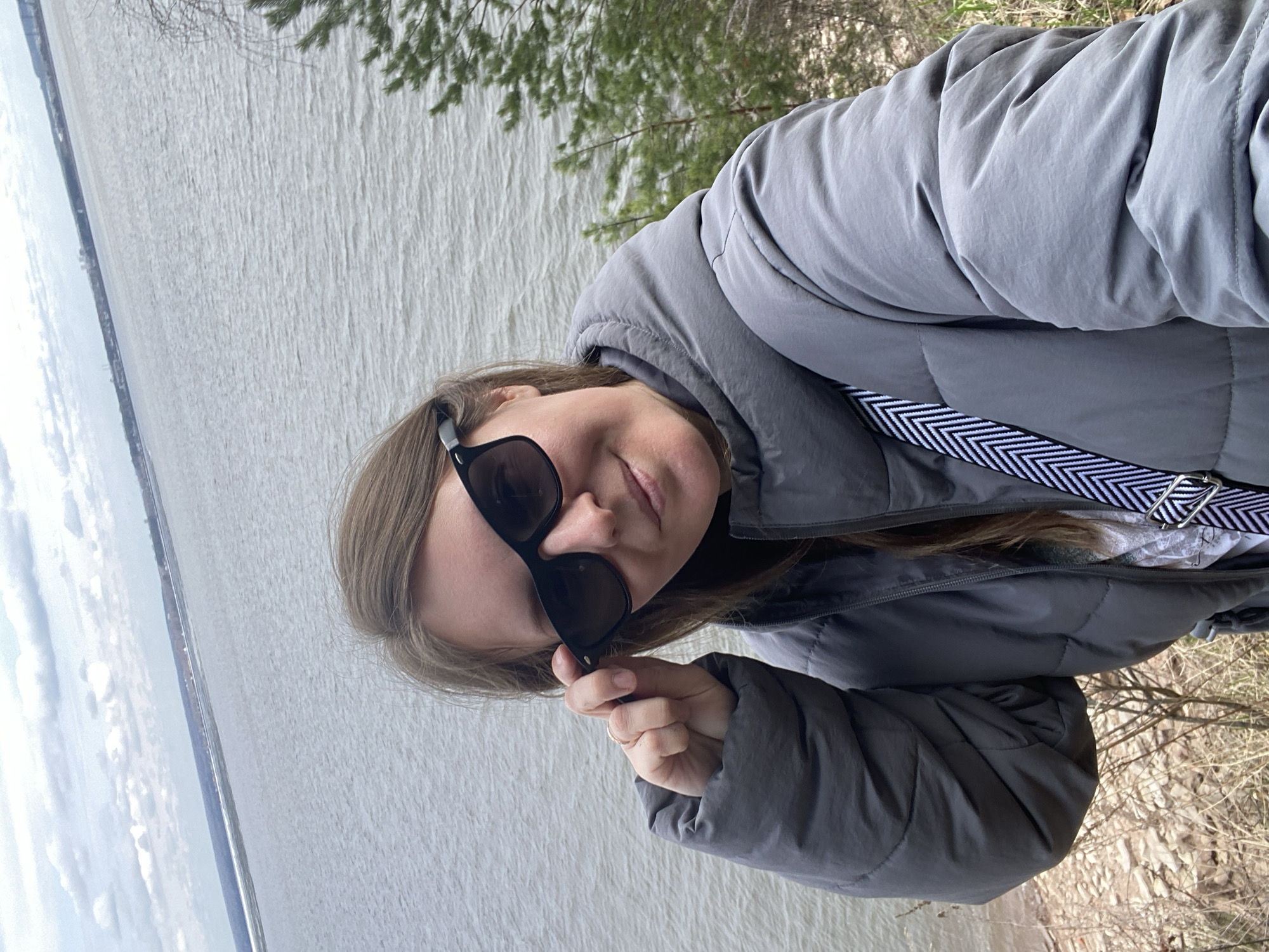 A woman with light brown hair wearing a grey puffer jacket and sunglasses stands outside with a body of water in the background. The woman is holding her sunglasses with her right hand. There are trees and greenery in the background. The location is Chaikovsky, Russia.
