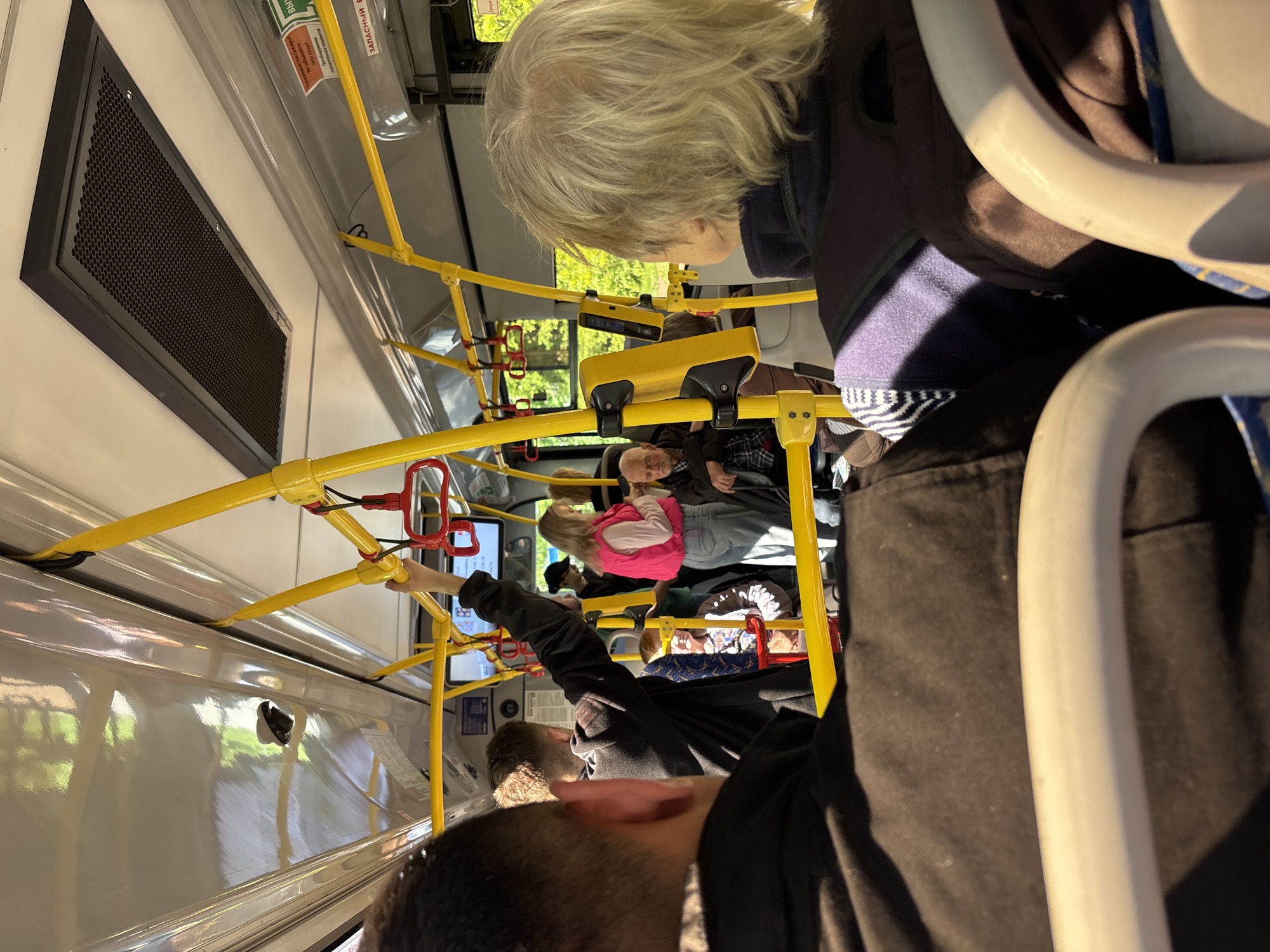 The image depicts the interior of a bus, likely a public transportation vehicle, filled with passengers. The bus is moving, as suggested by the background scenery visible through the windows. The scene is dominated by yellow handrails and poles that passengers use for support. Several people are visible, including an elderly woman with grey hair, a person wearing a pink vest, and others in various attire. One person is holding onto a handrail. The perspective is from a low angle, possibly from someone seated or standing near the front of the bus. Given the query parameters, the location is likely within the city of Vsevolozhsk, Russia, though the specific location is not visually discernible.