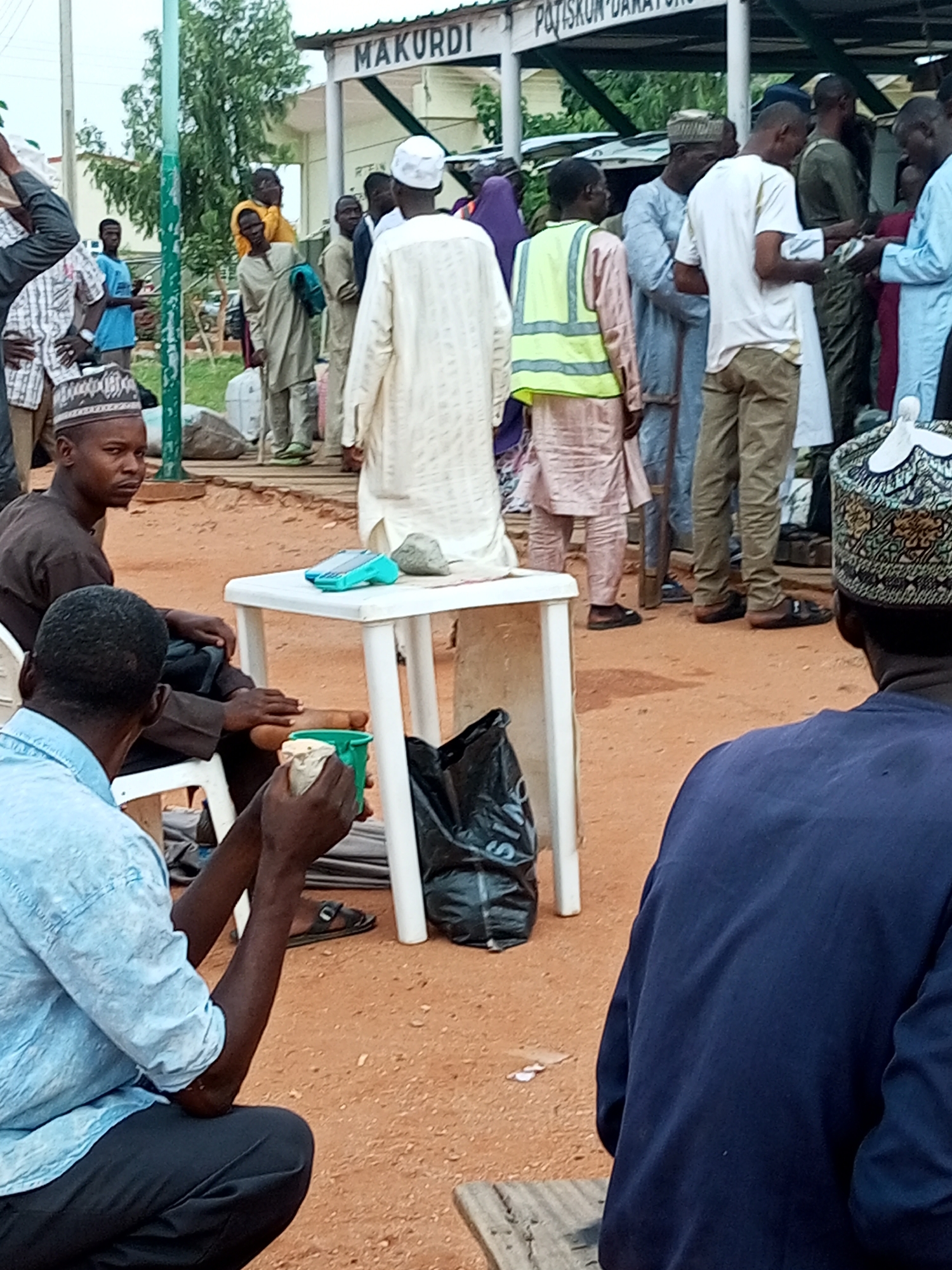 The image depicts an outdoor scene, likely in the city of Gombe, Nigeria, featuring a group of individuals gathered around a white plastic table. The background shows a building with signage that reads "MAKURDI" and "POTISKUM-DAKARTO." Several men, dressed in traditional attire and caps, are standing and interacting.
In the foreground, two men are seated. One man in a light blue shirt and dark trousers is looking away from the camera, with his arms resting on his lap. He appears to be holding a light-colored object in his right hand. The other seated man, in a dark brown tunic, is looking directly at the camera with a serious expression. He is seated in a white plastic chair.
On the white plastic table, a small green device, possibly a point-of-sale terminal, is visible along with a gray rock. Beside the table, a black plastic bag rests on the ground. The ground surface is covered in reddish-brown sand and small stones.
Other individuals are visible in the mid-ground and background. One man is wearing a bright yellow reflective vest over a traditional garment. Some people are holding what appear to be paper bills. The overall atmosphere suggests a public gathering or a transaction taking place. The presence of traditional clothing and the building signage hint at a local setting.