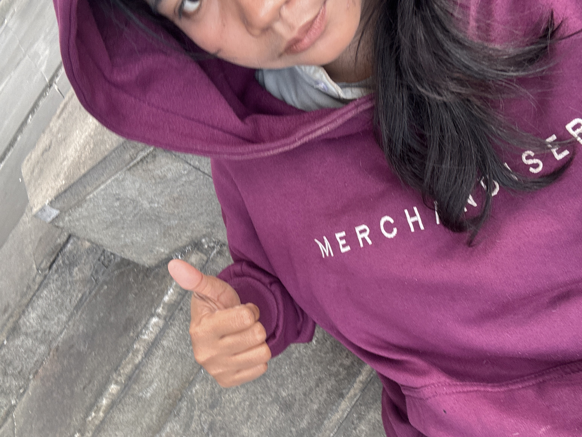 A woman's upper body is visible in this close-up, high-angle shot. She is wearing a maroon hoodie with white lettering on the front that appears to read "MERCHI..." and further down "SLR". Her right hand is raised with her thumb up in a "thumbs up" gesture. She has dark hair and her face is partially visible, looking towards the camera. The background consists of weathered concrete steps or tiered surfaces, typical of urban outdoor environments, possibly found in areas like the waterfront or parks in North Jakarta, Indonesia. The lighting suggests an overcast day.