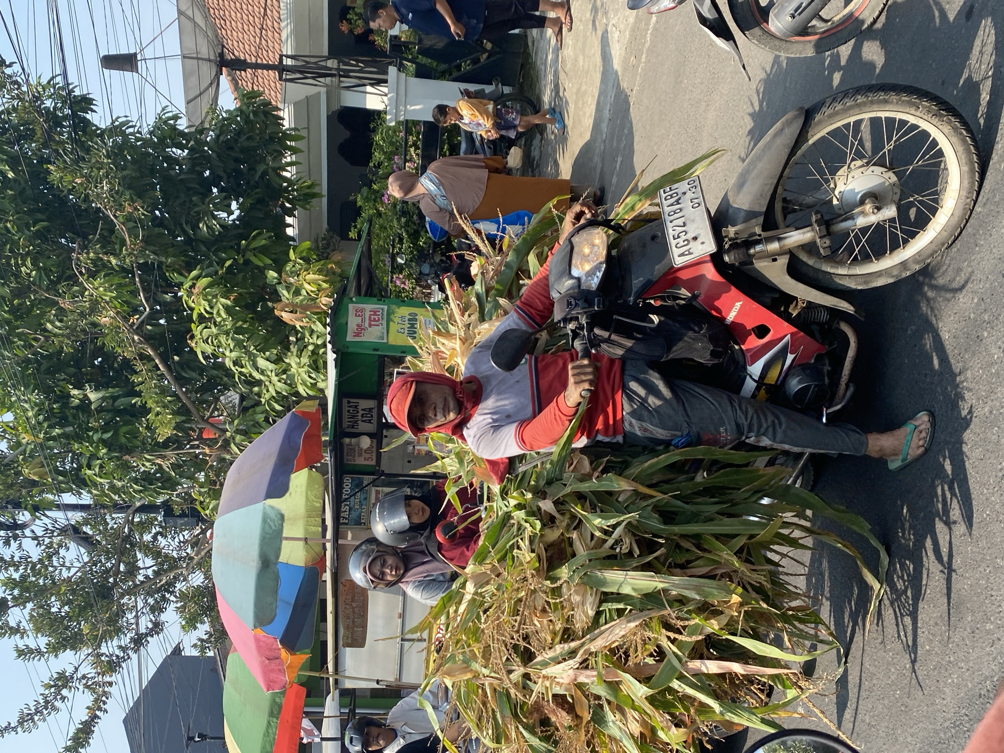 The image depicts a sunny street scene in the city of Kediri, Indonesia. A man wearing a red headscarf, red and white shirt, and grey pants is riding a red motorcycle heavily laden with a large quantity of corn stalks. He is looking forward, seemingly driving. Behind him, two women wearing helmets are also on the motorcycle, both looking forward. One woman wears a red top and the other a grey headscarf and red top. In the background, there are several colorful umbrellas, stalls, buildings, and trees. A satellite dish is visible atop a house in the distance. To the right, a man in a blue shirt and a young boy in a yellow shirt and shorts stand on the sidewalk, observing. The environment suggests a typical urban street with commercial activity and residential buildings.