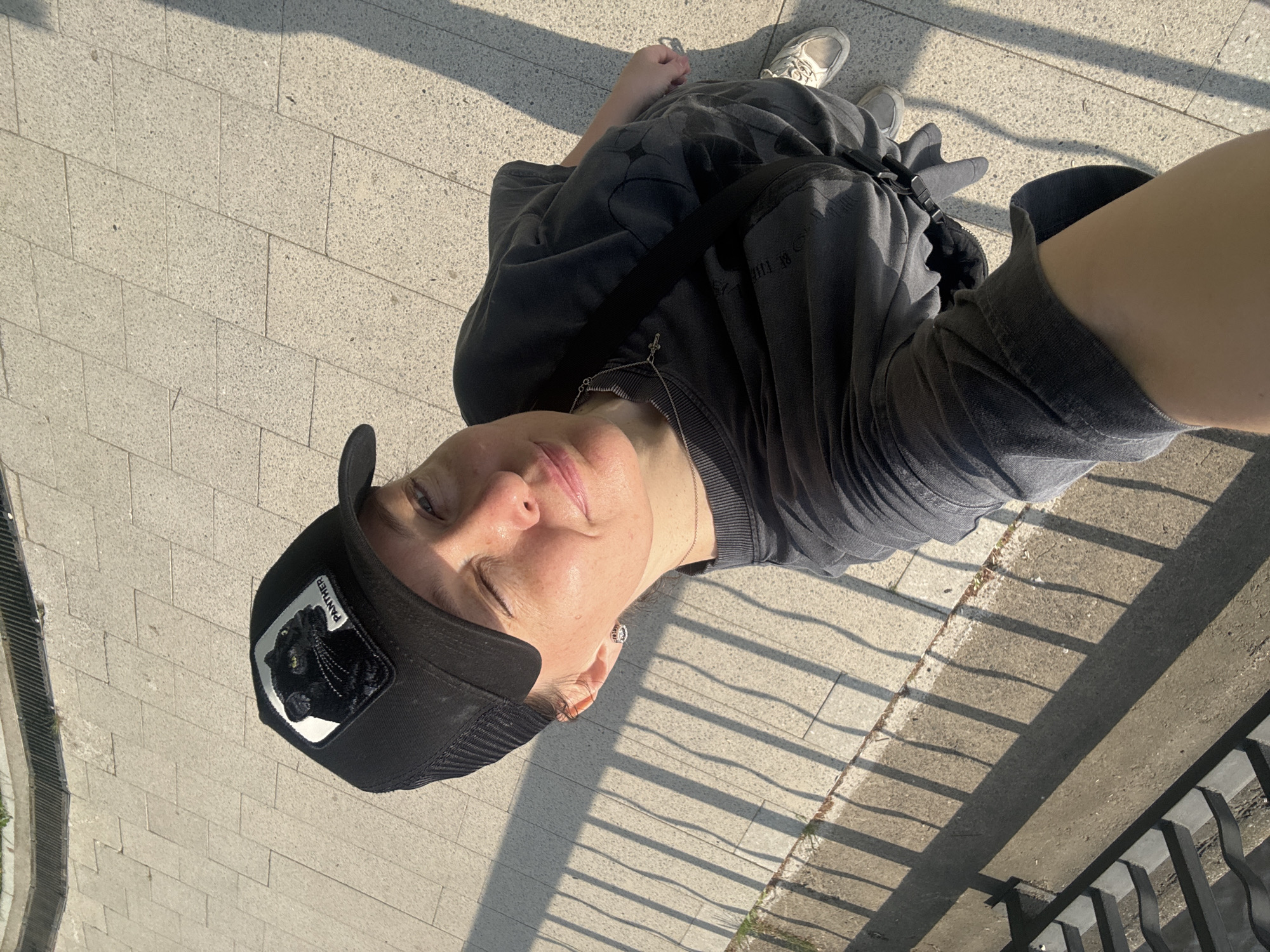 A person takes a selfie in Chaikovsky, Russia. The person is wearing a black cap with a black panther logo on the front, a gray t-shirt, and a cross necklace. The person is lying on a paved surface. Sunlight is casting shadows from a nearby railing.