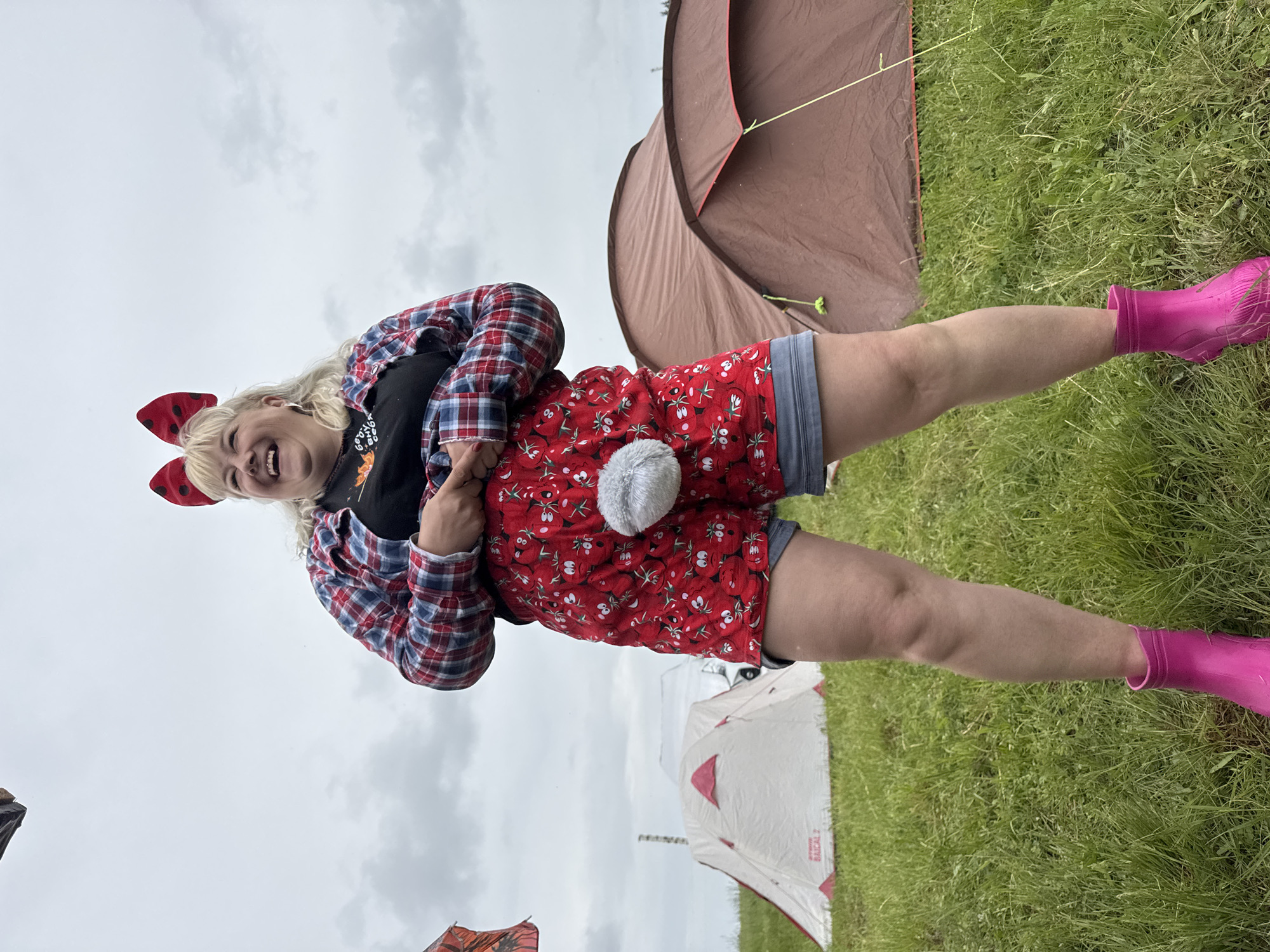A joyful blonde-haired individual stands upright in a grassy outdoor setting, likely a camping ground or festival, given the tents in the background. The person is smiling broadly with an open mouth, conveying a sense of enthusiasm and fun. They are wearing a red and black plaid long-sleeved shirt over a black t-shirt. Their lower body is adorned with playful red shorts featuring a repeating pattern of white-outlined, smiling fruit or animal faces, complete with a fluffy grey pom-pom tail on the backside. A pair of vibrant, almost fluorescent pink rain boots cover their feet, suitable for the potentially wet and grassy environment. A large red bow with white polka dots, resembling a ladybug, is worn in their hair.
In the background, two tents are visible. To the right, a brown pop-up tent is partially pitched. To the left, further back, another tent is visible, predominantly white with red accents, and the text "RADICAL 2" can be discerned on its side. The ground is covered in green grass. The sky is overcast with grey clouds, indicating a daytime scene. The overall atmosphere suggests an outdoor event or casual gathering in Большая Кивара, Russia, characterized by a playful and happy mood.