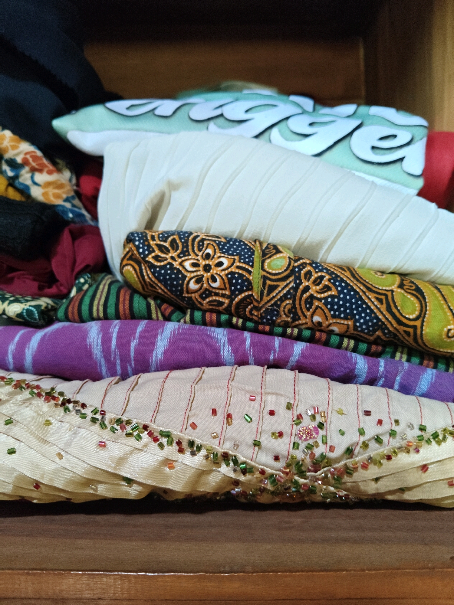 The image shows a stack of folded textiles, likely fabrics or clothing, resting on a wooden surface, possibly a shelf or drawer in a dresser. The textiles are layered and vary in color and pattern. The topmost item appears to be a light green fabric with a white wave-like pattern. Beneath this is a cream-colored pleated fabric. The next layer is a fabric with a dark blue base and an intricate orange, yellow, and green floral or paisley pattern. Below that is a purple fabric with thin, light blue vertical stripes. The bottom visible textile is a light beige fabric with red vertical stripes and a decorative border of small, colorful beads in shades of red, green, and yellow. The overall impression is of a neatly stored collection of textiles. The specific location of Sumberwono, Indonesia is not depicted or inferable from the visual information provided in the image.