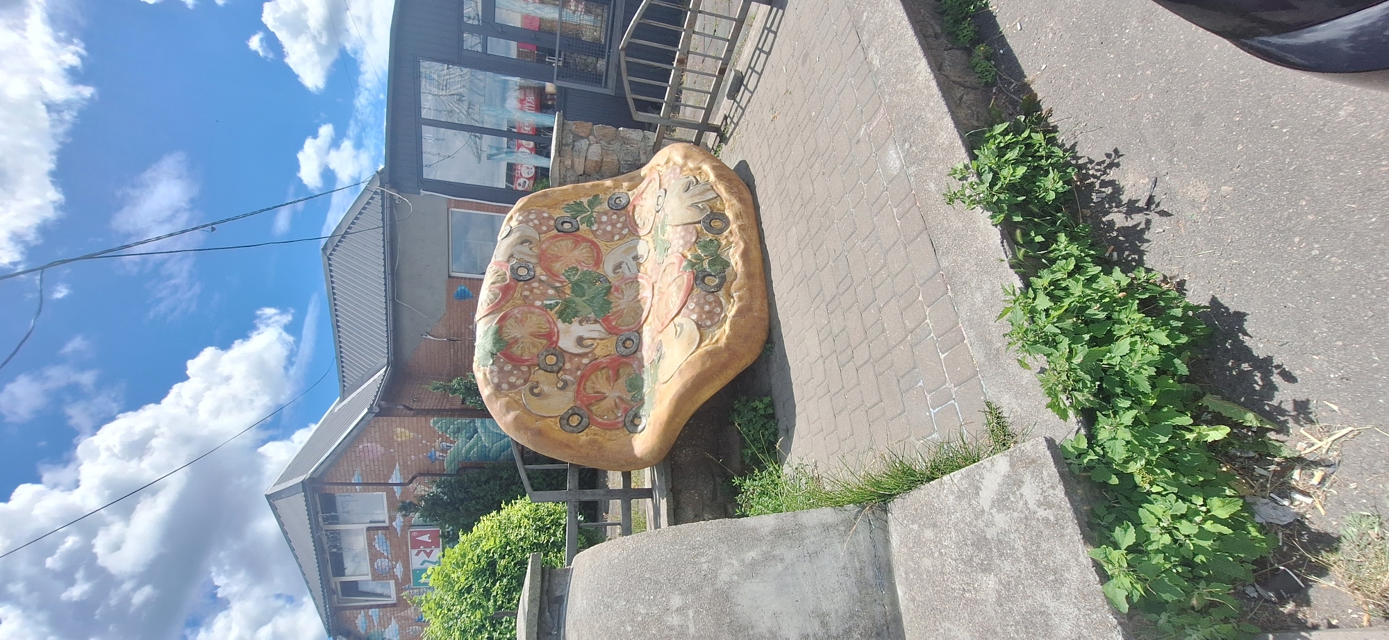 The image shows a unique outdoor seating area outside a building, likely a restaurant or cafe, in Bila Tserkva, Ukraine. The centerpiece is a large, concrete bench sculpted to resemble a slice of pizza, complete with toppings like mushrooms, tomatoes, and olives. This pizza-shaped bench sits on a paved area, bordered by a low stone wall and a metal fence. Behind the bench, the building has a brick facade adorned with painted murals depicting clouds, a cityscape, and geometric shapes. The building features a gray, sloped roof and large windows. Greenery, including bushes and grass, surrounds the seating area and grows along the edges of the pavement and sidewalk. The sky above is blue with scattered white clouds. The overall impression is a quirky and inviting public space.