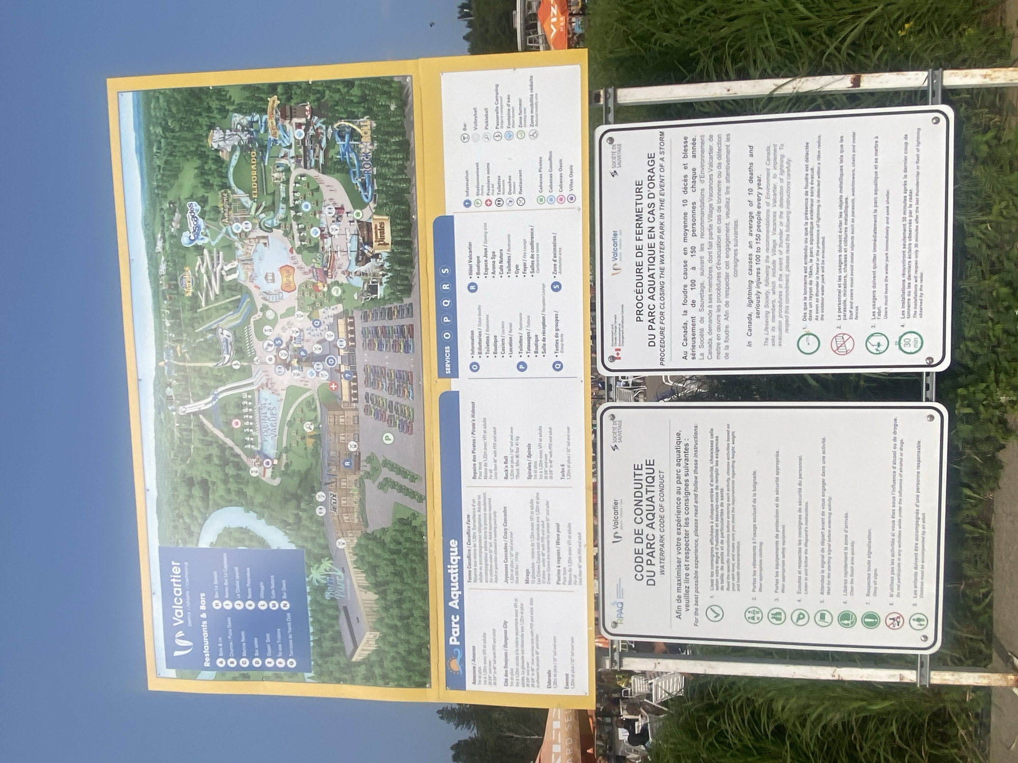 The image shows a clear, bright day at the Valcartier Vacation Village, located in Saint-Gabriel-de-Valcartier, Canada. A large, yellow sign dominates the upper portion of the frame, displaying a detailed map of the water park. The map highlights various attractions, restaurants, and services within the park, including areas labeled "Amazonia," "Eldorado," "Pirates," and "Rock 'n Roll." Below the main map, two additional signs are visible. The sign on the left, titled "CODE DE CONDUITE DU PARC AQUATIQUE," outlines rules and regulations for park visitors. The sign on the right, "PROCÉDURE DE FERMETURE DU PARC AQUATIQUE EN CAS D'ORAGE," provides instructions for closing the park in the event of a thunderstorm. The environment appears to be lush and green, with trees and vegetation surrounding the park structures. In the foreground, a grassy area with some chairs is partially visible, suggesting a seating or relaxation zone.