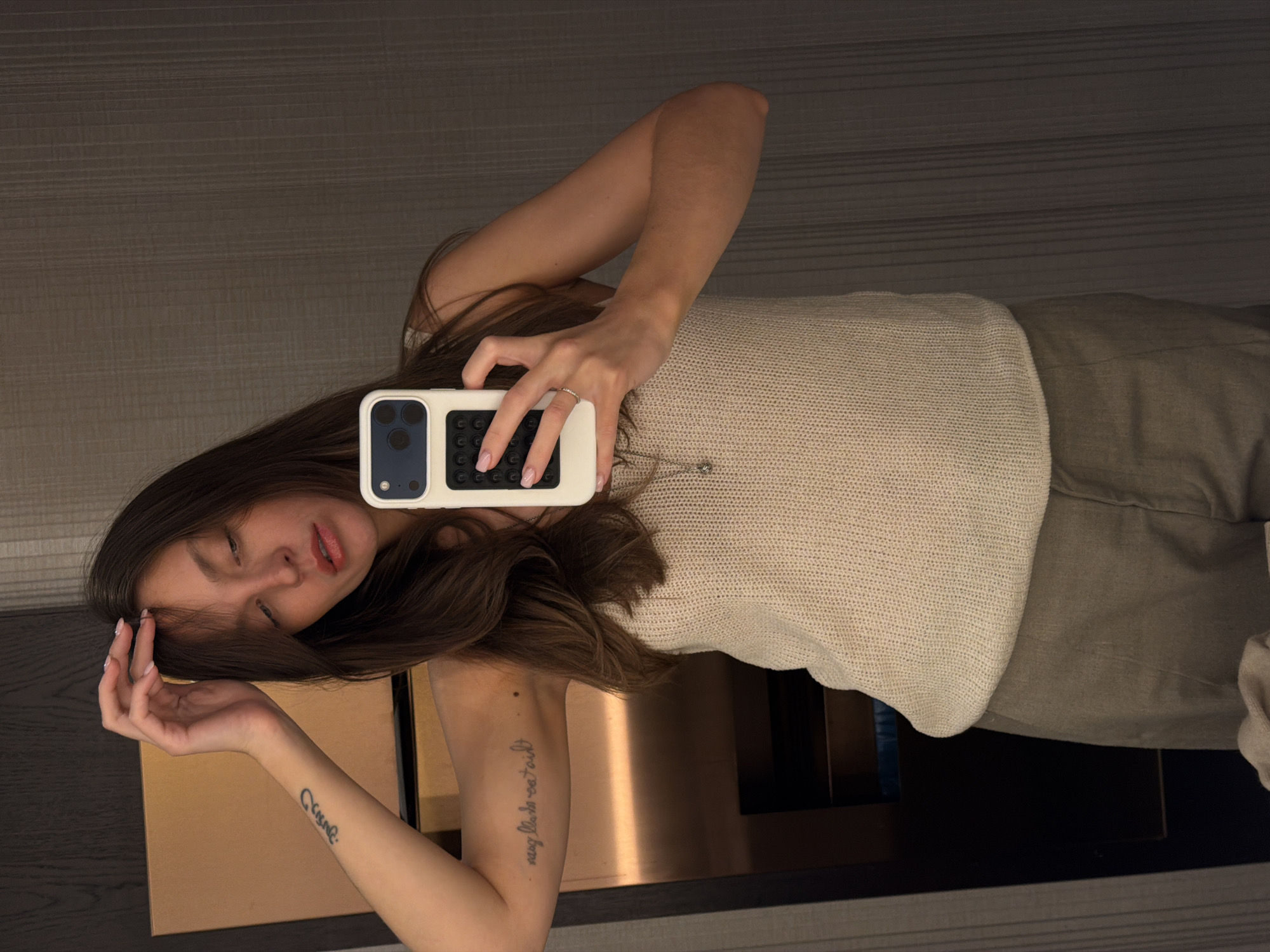 A woman takes a mirror selfie indoors. She is wearing a sleeveless light beige top and light brown trousers. She holds a smartphone with a white and black case in her right hand. The mirror reflects a cabinet with a glossy copper-colored surface. The environment suggests a modern interior, possibly a hotel room or apartment. There is no indication the photograph was taken in Budva, Montenegro.