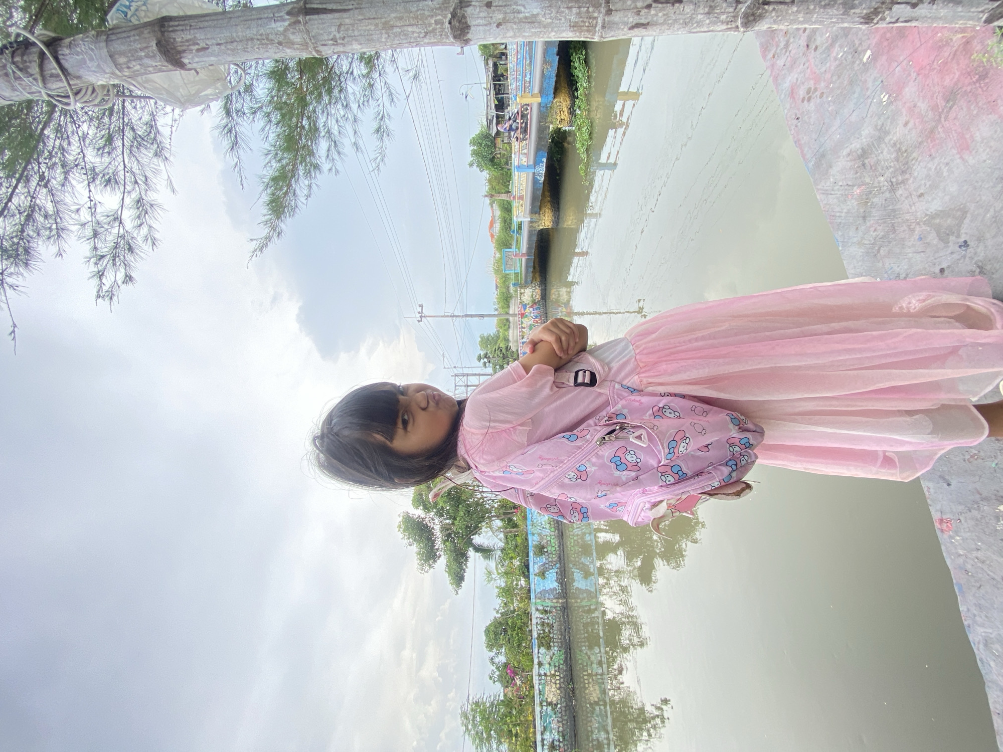 A child wearing a pink dress and a backpack stands next to a body of water, likely a canal or river, in Kalitengah, Indonesia. The water surface reflects the sky and surrounding vegetation. A bridge or structure is visible in the background. The scene is overcast. The child appears to be looking off to the side.