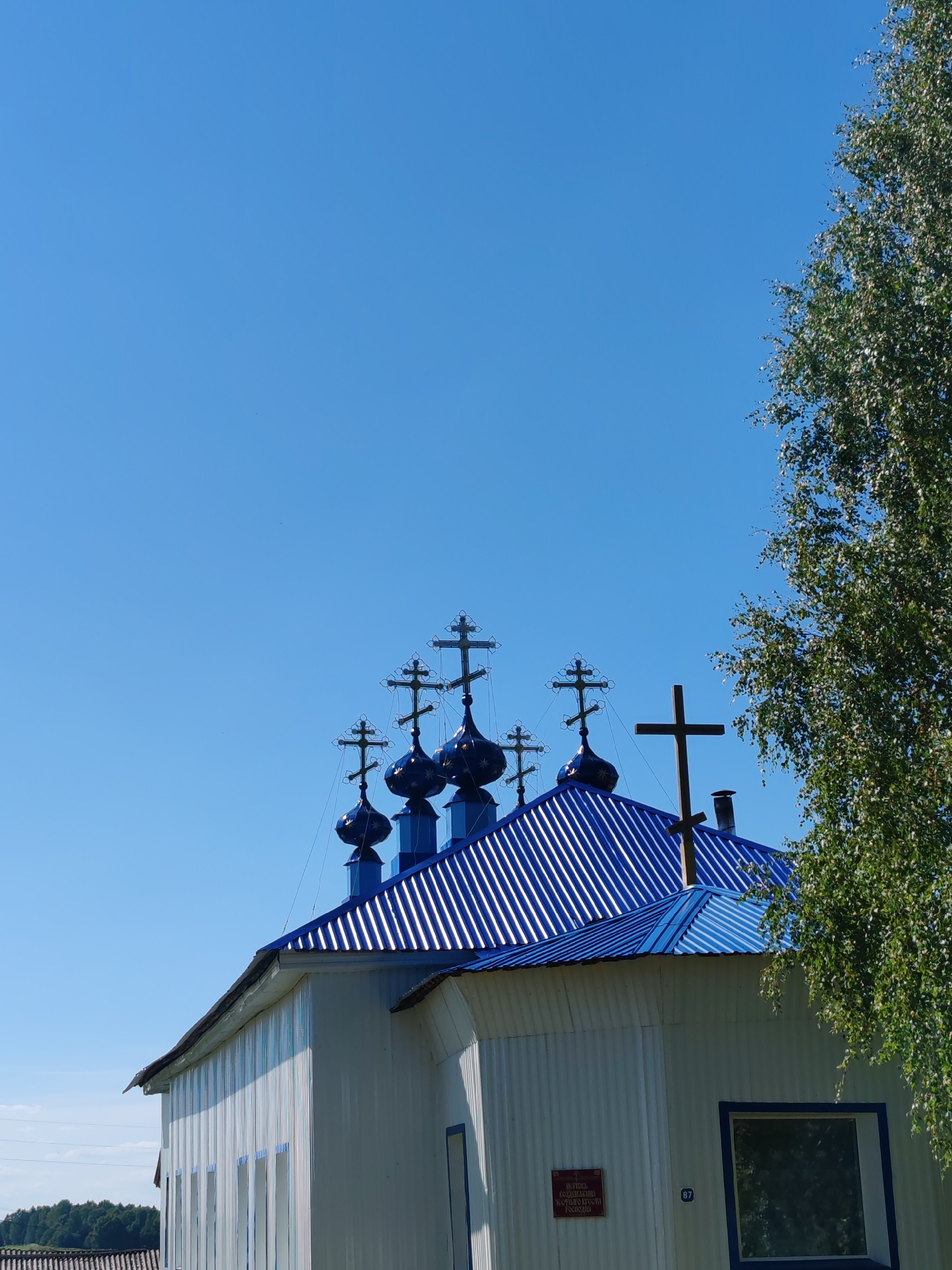 A low-angle, full shot captures the top portion of a white building with a corrugated metal exterior, featuring a bright blue, corrugated metal roof. Several blue domes, each topped with a multi-barred cross, rise from the roof. A single wooden cross stands on the roofline to the right of the domes. A large birch tree with green leaves occupies the right side of the frame, partially obscuring the sky. The sky is a clear, bright blue. The building appears to be a church, possibly the Church of the Intercession of the Blessed Virgin Mary, located in the village of Chukhlom, Kostroma Oblast, Russia.