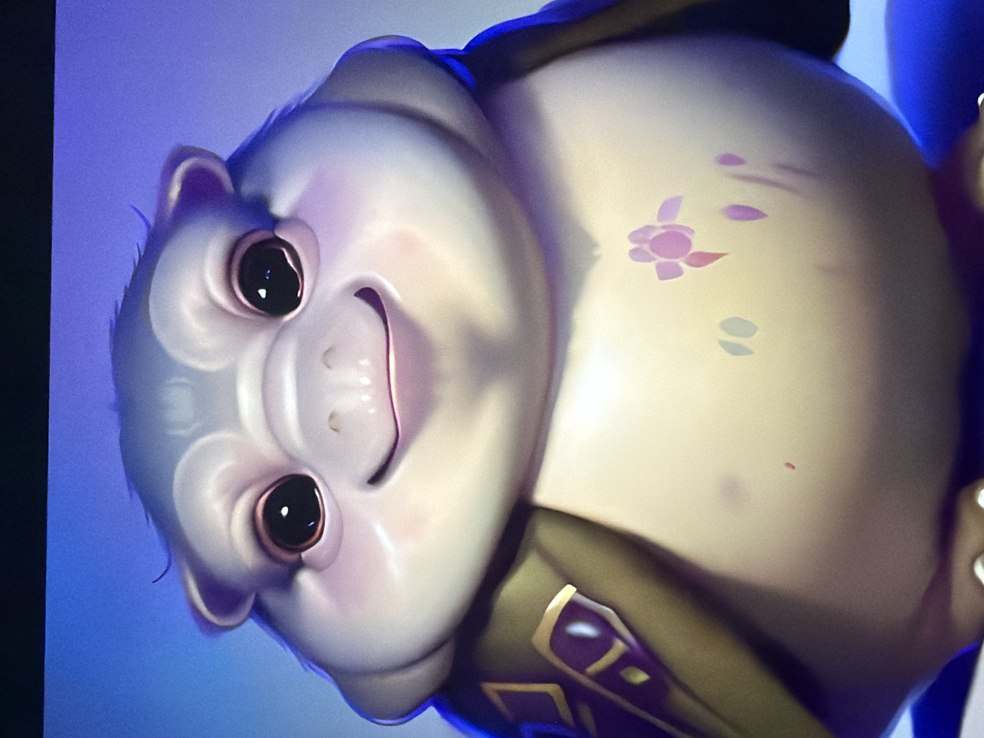 The image is a close-up, digital rendering of a cartoonish, grey, and plump creature. The creature has large, dark eyes with bright reflections, small ears, a wide smile, and rosy cheeks. It is wearing a green garment with purple and gold embellishments on its left side. The creature's chest is adorned with a pink floral pattern and some abstract marks. The background is a gradient of blue. There is no indication of a location in Kyiv, Ukraine, or any real-world environment.