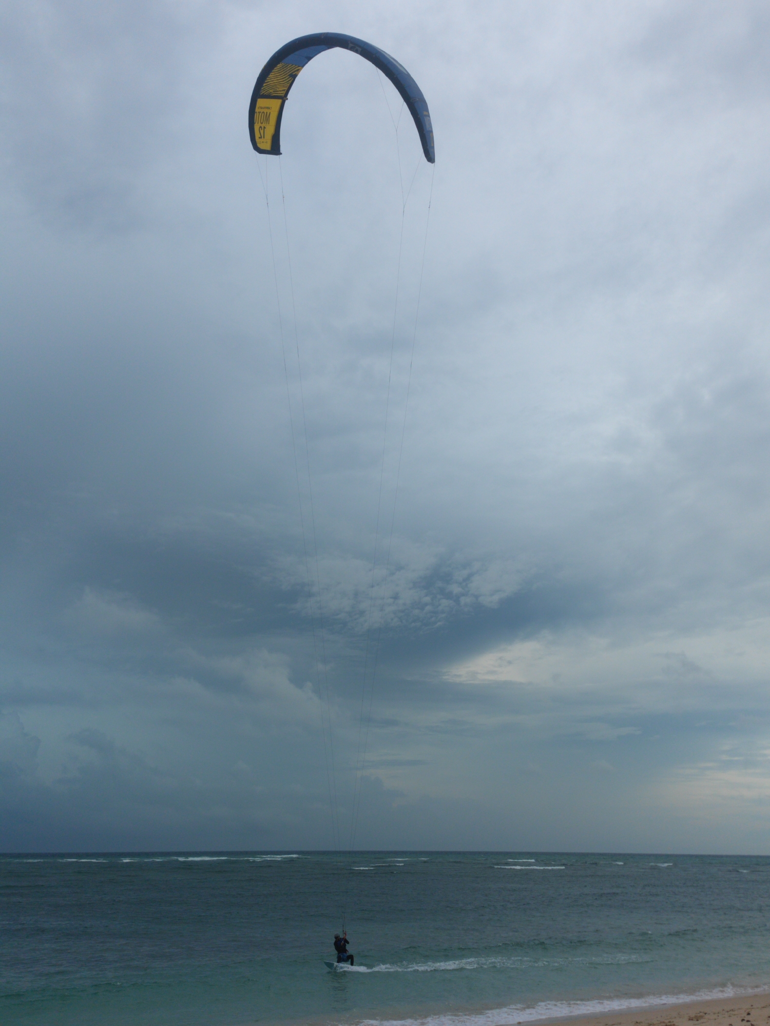 The image depicts a kitesurfer actively engaged in the sport on a body of water under a heavily overcast sky. The scene is set in a coastal area, likely Phú Quý Special Administrative Region, Vietnam.
In the mid-ground, a single person, seen as a dark silhouette, stands on a small board in the water, generating a white wake. They are connected by multiple thin lines to a large, arched kitesurfing kite positioned high in the upper center of the frame. The kite is predominantly dark blue with a striking yellow segment near its center. Clearly visible text on the yellow section of the kite, oriented upside down, reads "TOM ST". The word "NORTH" is also faintly discernible in white on the blue part of the kite.
The sky occupies the majority of the upper frame, filled with thick, dark grey clouds that create a diffused, subdued light. This suggests an overcast day, possibly indicating impending rain or a generally cloudy weather condition. Below the horizon line, the ocean extends, showing gentle white-capped waves further out and smaller ripples closer to the shore. The water near the person is a clear turquoise-green, gradually deepening to a darker blue-green further away. A narrow strip of sandy beach is visible in the bottom right corner of the image. The overall impression is one of determined activity against a dramatic, moody sky.