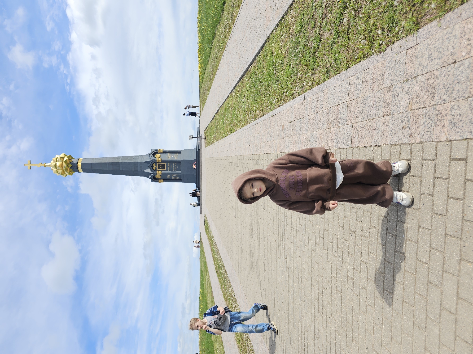 The image captures a scene along a brick-paved walkway in Unknown, Unknown. A child wearing a brown hoodie and matching pants stands on the walkway. In the background, a tall, dark monument with a golden dome and cross on top is visible against a blue sky with scattered clouds. Several people are seen near the monument. A man wearing jeans, a blue checkered shirt, and a gray t-shirt with a black bag slung across his chest is walking along the walkway in the lower left corner. There are strips of green grass along the sides of the walkway. The lighting suggests it is daytime.