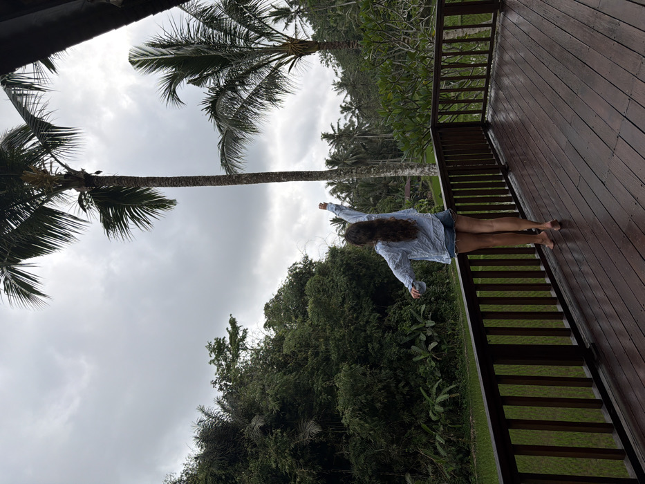 A single female subject stands on a dark brown wooden deck, viewed from behind, in Tegal Suci, Indonesia. She is barefoot, wearing a light blue long-sleeved shirt and denim shorts. Her long, wavy brown hair extends down her back. Her right arm is raised vertically and extended upwards, while her left arm is slightly bent and lowered. The wooden deck, which appears wet, is surrounded by a matching dark brown wooden railing. Beyond the railing, a lush tropical landscape unfolds under an overcast grey sky. This environment features dense green foliage, numerous tall palm trees, and a vibrant green rice paddy field visible in the mid-ground to the left. A dark wooden structural element is partially visible in the upper right corner of the frame.