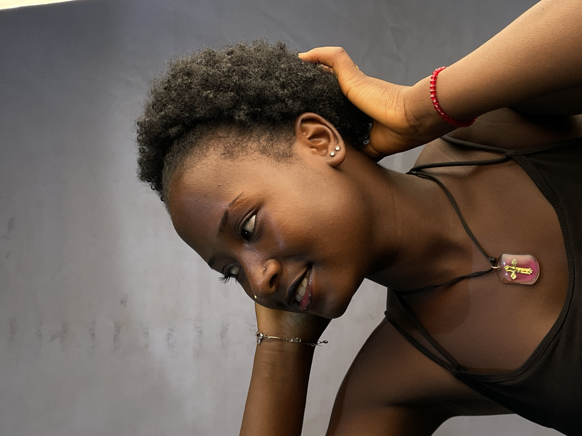 A medium shot of a young, dark-skinned woman with short, curly afro-textured hair, wearing a black tank top. She is looking down and to the left. Her left hand supports her head, and her right hand touches the back of her head. She has a nose piercing and is wearing earrings, a delicate bracelet on her left wrist, a red beaded bracelet on her right wrist, and a necklace with a pendant featuring a golden cross. The background is a mottled gray. No information confirms the location. The scene is likely indoors, given the controlled lighting and lack of environmental elements. No action or interaction is readily apparent.