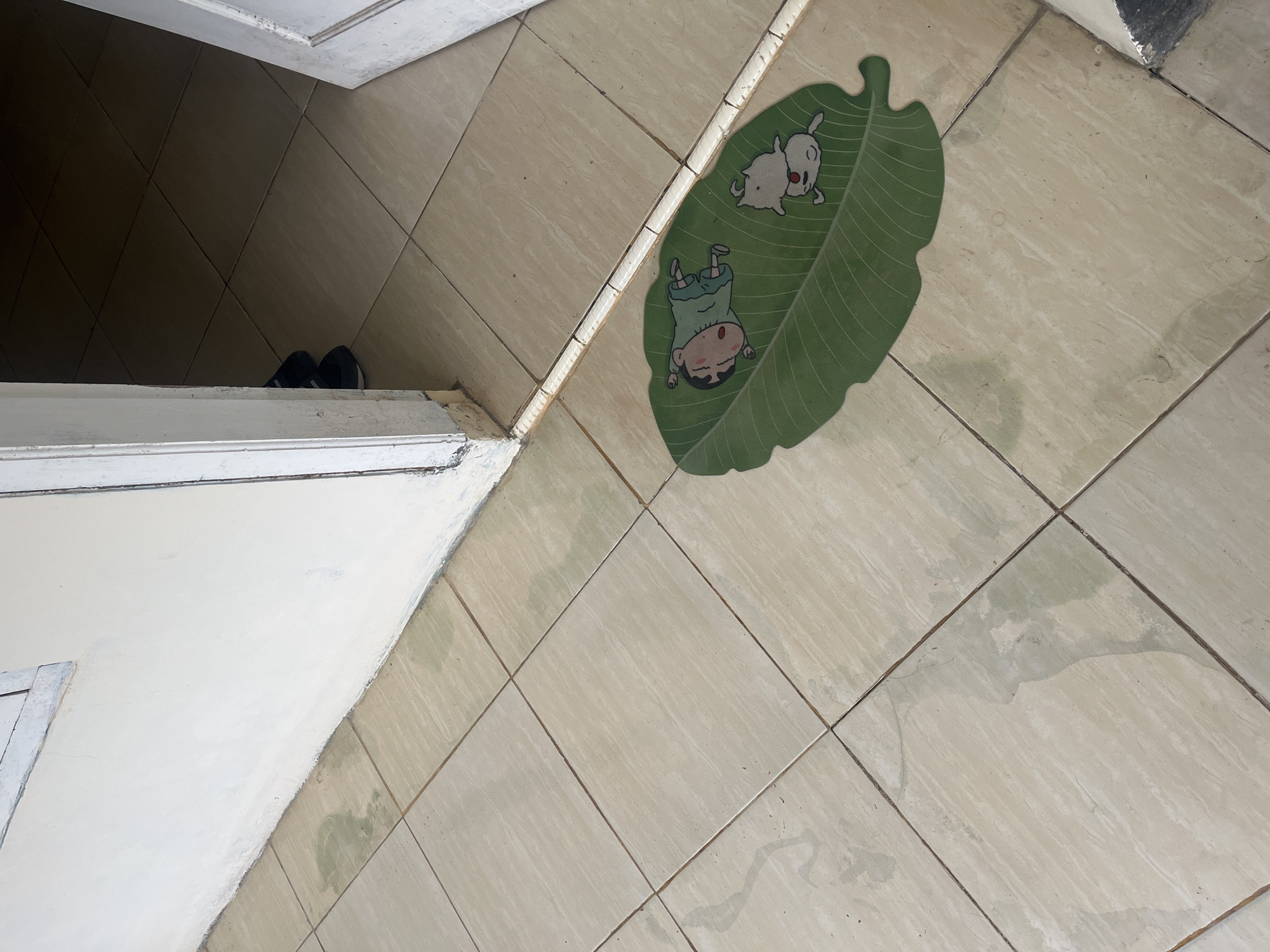 A doormat featuring a cartoon character and a dog is placed on a tiled floor near the entrance of a building. The doormat is green and shaped like a leaf. The cartoon character, resembling Shin-chan, is lying down on the leaf. A small white dog is also on the leaf, next to the character. The floor tiles are beige and have a patterned texture. There are some water stains on the tiles, particularly on the left side of the image. A white wall and a doorway are visible in the background, with a pair of dark sandals at the entrance. The overall scene appears to be an outdoor or semi-outdoor area, possibly a porch or patio. The image provides a close-up view of the doormat and its surroundings, with the building's entrance framing the scene. The lighting suggests it is daytime.