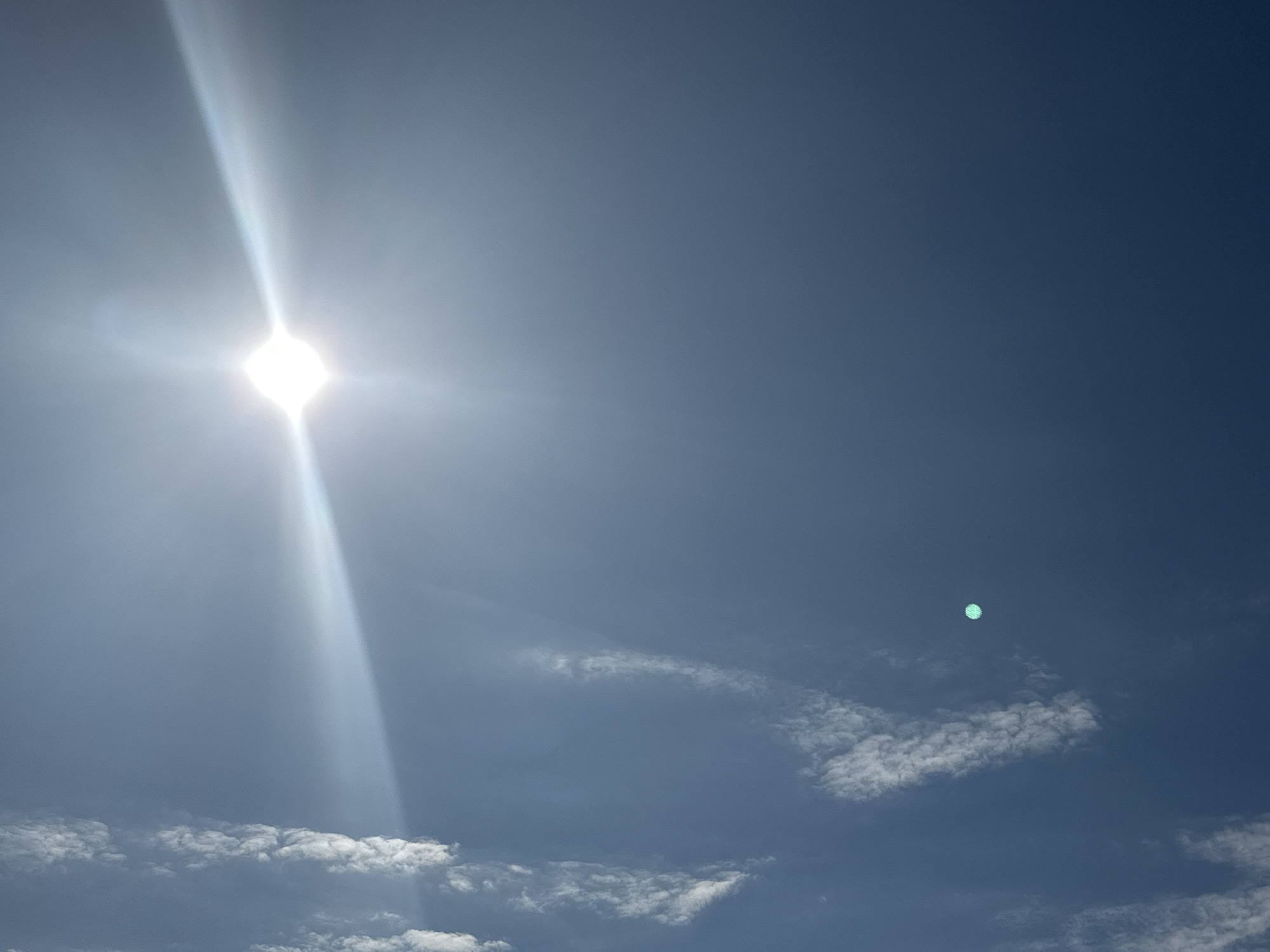 The image captures a bright sun casting a prominent lens flare across a clear, deep blue sky. A few scattered white clouds are visible in the lower portion of the frame, with more defined cloud formations forming a band near the bottom. A small, luminous green circular object appears in the upper right quadrant of the sky, possibly a reflection or atmospheric phenomenon. No specific landmarks or urban features are discernible in this image, therefore its location in Podolsk, Russia cannot be confirmed.