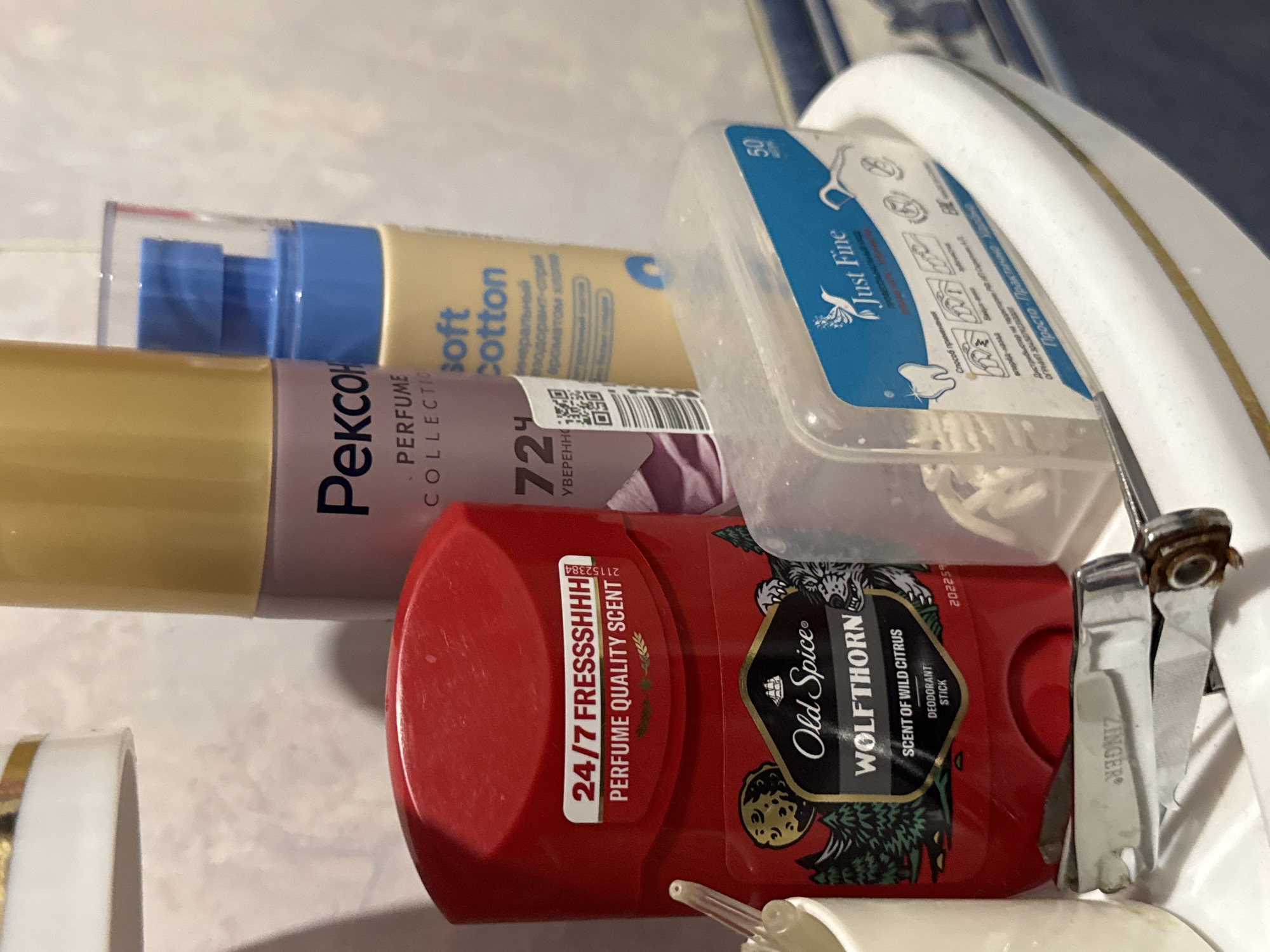 The image shows a close-up of a collection of personal care products and grooming tools on the edge of a white ceramic sink or countertop, likely in a bathroom. The objects are arranged haphazardly, suggesting they are in frequent use. Dominating the foreground is a red Old Spice "Wolfthorn" deodorant stick, with a label indicating "PERFUME QUALITY SCENT" and "DEODORANT STICK." Below it are a pair of metallic nail clippers with the brand name "ZINGER" visible. To the right of the deodorant stick, a clear plastic container holds what appears to be dental floss or interdental brushes, with a blue label that reads "Just Fine" and includes diagrams of how to use the product. Behind these items, several cylindrical bottles and containers are partially visible. One is a tall, slender bottle with a gold cap and a pinkish-purple body, featuring the brand name "Рексон" (Rekson) and text that partially reads "PERFUME COLLECTION." Next to it is a smaller, pale yellow bottle labeled "soft Cotton." The background is a plain, light-colored textured surface. There are no people or animals present in the image. The lighting is soft, indicating an indoor setting, possibly with artificial light. The overall impression is one of everyday domestic use of toiletries. The location is described as Яромінскі сельскі Савет, Belarus, suggesting this is a bathroom in a residential setting in that area.