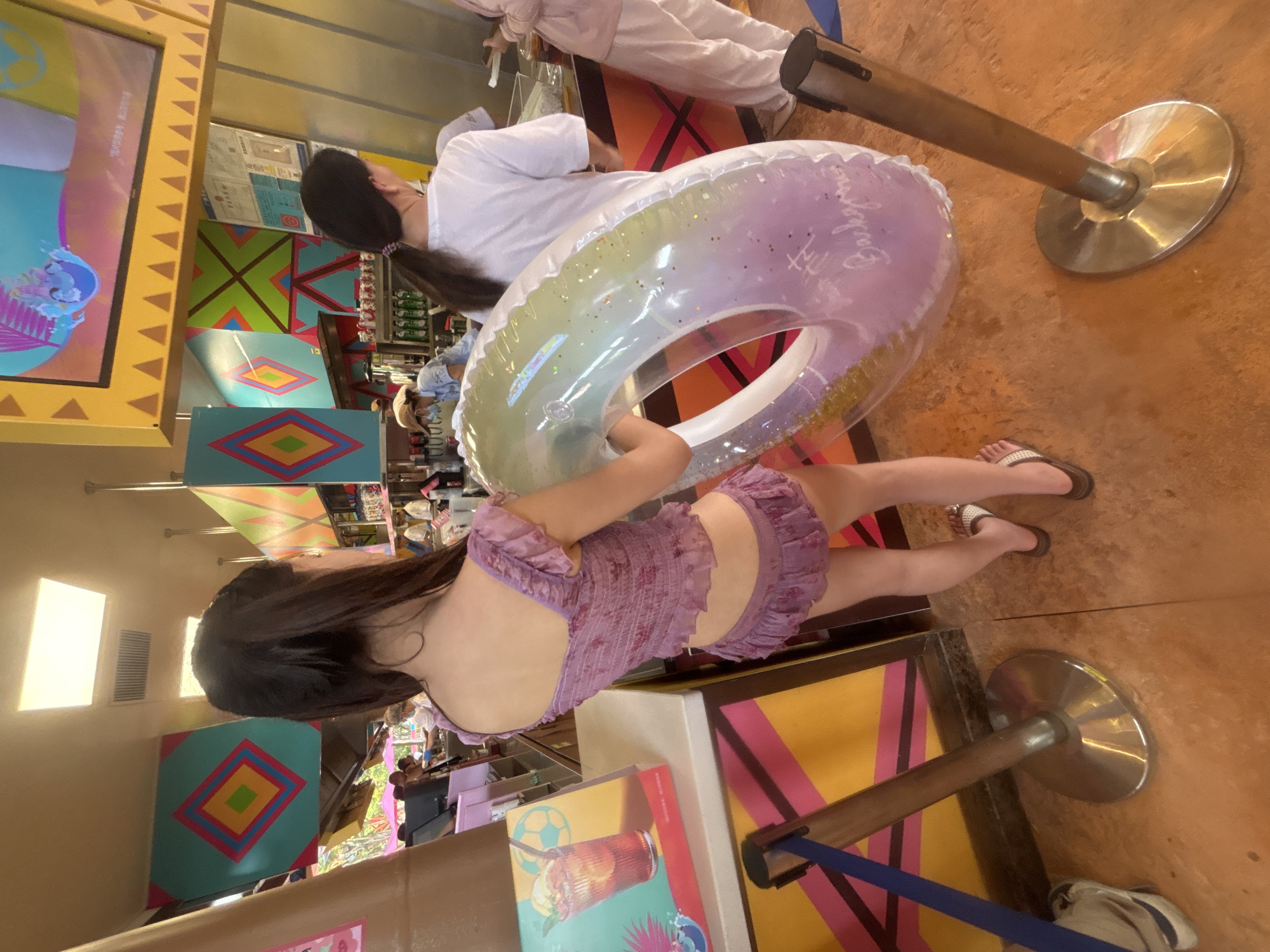 In Panyu District, China, a young woman, seen from behind, stands in what appears to be an indoor concession area, possibly within an amusement park or water park. She is wearing a purple, floral-patterned bikini set with ruffled details. In her right arm, she holds a large, translucent inflatable ring with iridescent hues and glitter. The background reveals a food or beverage counter with several people in the process of ordering or being served. Colorful, geometric decorations adorn the walls and ceiling. A digital screen displaying an animated character hangs above the counter. The floor is a textured orange-brown surface, and stanchions with blue ropes mark a queue or pathway. A backpack is visible on the floor to the left.