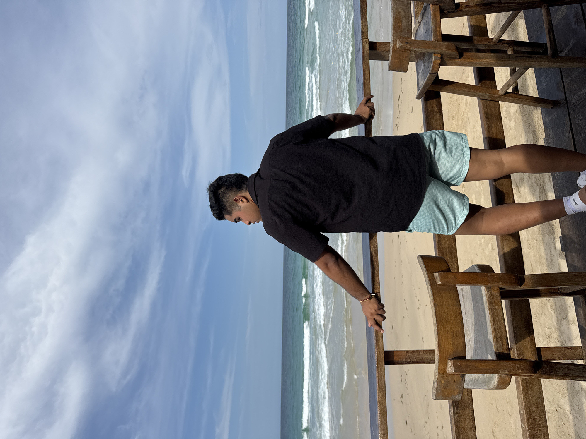 The image depicts a serene coastal scene in El Tirano, Venezuela, centered on a man observing the ocean from a wooden deck. The man, with a medium-brown complexion and short, dark hair, is dressed in a black short-sleeved shirt, light blue and white patterned shorts, white athletic shoes, and white socks, which bear a small, unreadable black logo on the right sock. He wears a golden-colored bracelet on his right wrist. He stands in profile, leaning slightly forward with both hands resting on a sturdy wooden railing that forms the edge of the deck.
The background showcases the calm, blue-green waters of the ocean, with gentle white waves breaking near a light-colored sandy shore. Above, the sky is a soft blue, adorned with wispy white clouds, suggesting clear and pleasant weather. The bright daylight, likely late morning or early afternoon, casts subtle shadows from the wooden structures on the deck. Behind the man and further into the deck area, several rustic wooden chairs are visible. The overall composition conveys a sense of peaceful observation and relaxation by the sea.