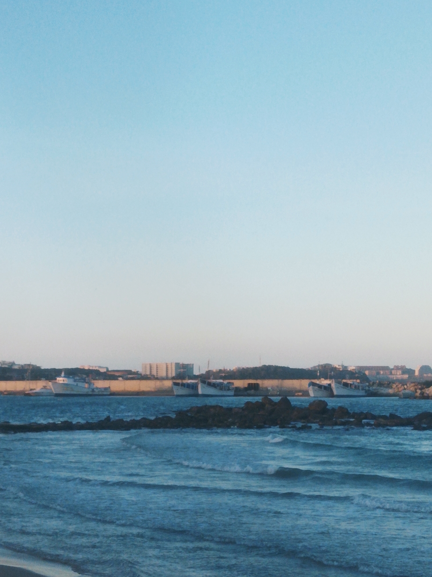 The image captures a coastal scene with a light blue sky dominating the upper portion. Below the sky, the sea stretches across the lower half of the frame, with visible waves. In the middle ground, there is a line of dark rocks extending across the water. Behind these rocks, closer to the horizon, there is a land mass with buildings and structures visible. There are also several ships docked or anchored near the coast. The image appears to be taken at or near Playa Moreno in Porlamar, Venezuela.